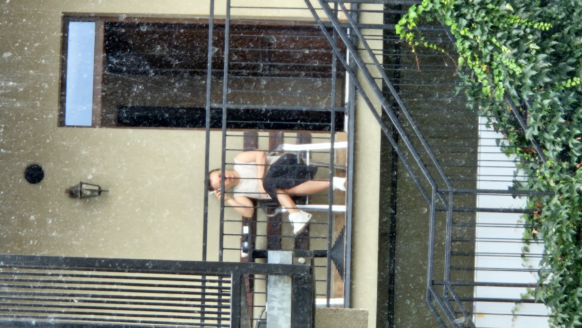 A woman is seated on a chair on a balcony, smoking a cigarette. She is wearing a light-colored, sleeveless top and a dark skirt. Her legs are crossed, and she is wearing white sneakers. The balcony has a black metal railing, and behind her is a closed door with a window above it. The building's exterior wall is a light beige color. It appears to be raining, as evidenced by water droplets on the glass of the door and on the overall image, creating a blurred effect. The balcony is on the second story of the building. Below the balcony, there is a metal staircase with a black railing, partially covered by green ivy. The scene is viewed from below and to the side, with a vertical black metal structure on the left side of the image. The overall impression is of an urban setting, possibly a residential building.