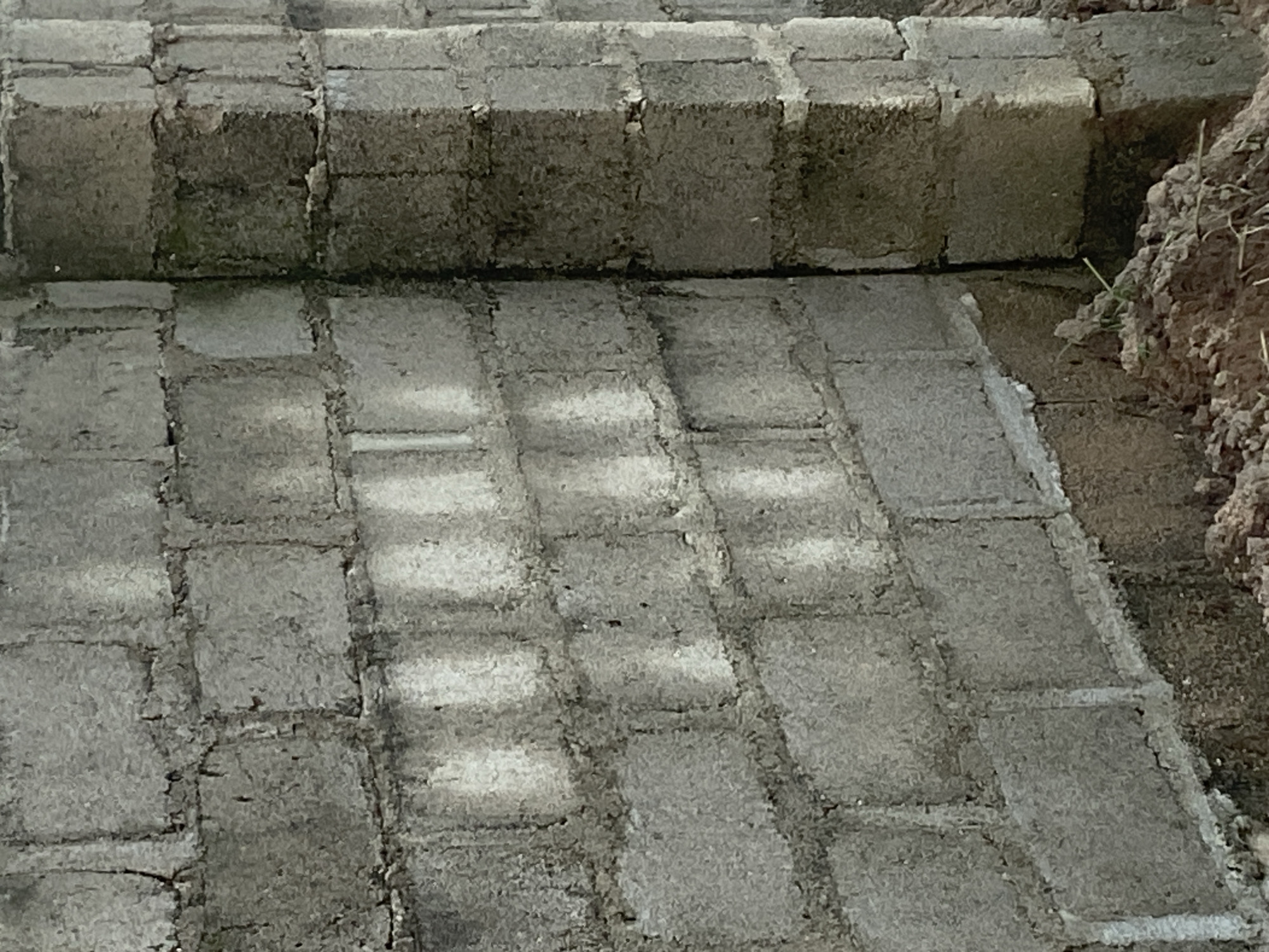 The image depicts a section of an unfinished cinder block wall and an adjacent pillar, likely part of a construction site in Jauro Yinu, Nigeria. The wall is constructed from rectangular cinder blocks laid in a staggered pattern. Some blocks show signs of wear and tear, with chipped edges and rough surfaces. Light and shadow play across the wall, highlighting the texture of the blocks and the mortar lines between them. Vertical, lighter-colored streaks are visible on the wall, suggesting where water may have run down.
The pillar, positioned at the corner of the wall, is also made of cinder blocks and appears to be of a more robust construction. It exhibits darker staining and a rougher texture than the wall. The base of the wall and pillar is surrounded by loose soil and small mounds of dirt, indicating that the ground level has not yet been finalized. Scattered blades of grass are visible amongst the soil. The scene is devoid of any people or specific identifiable objects other than the construction materials. The overall impression is one of ongoing construction in an outdoor, rural or semi-rural setting within Jauro Yinu.