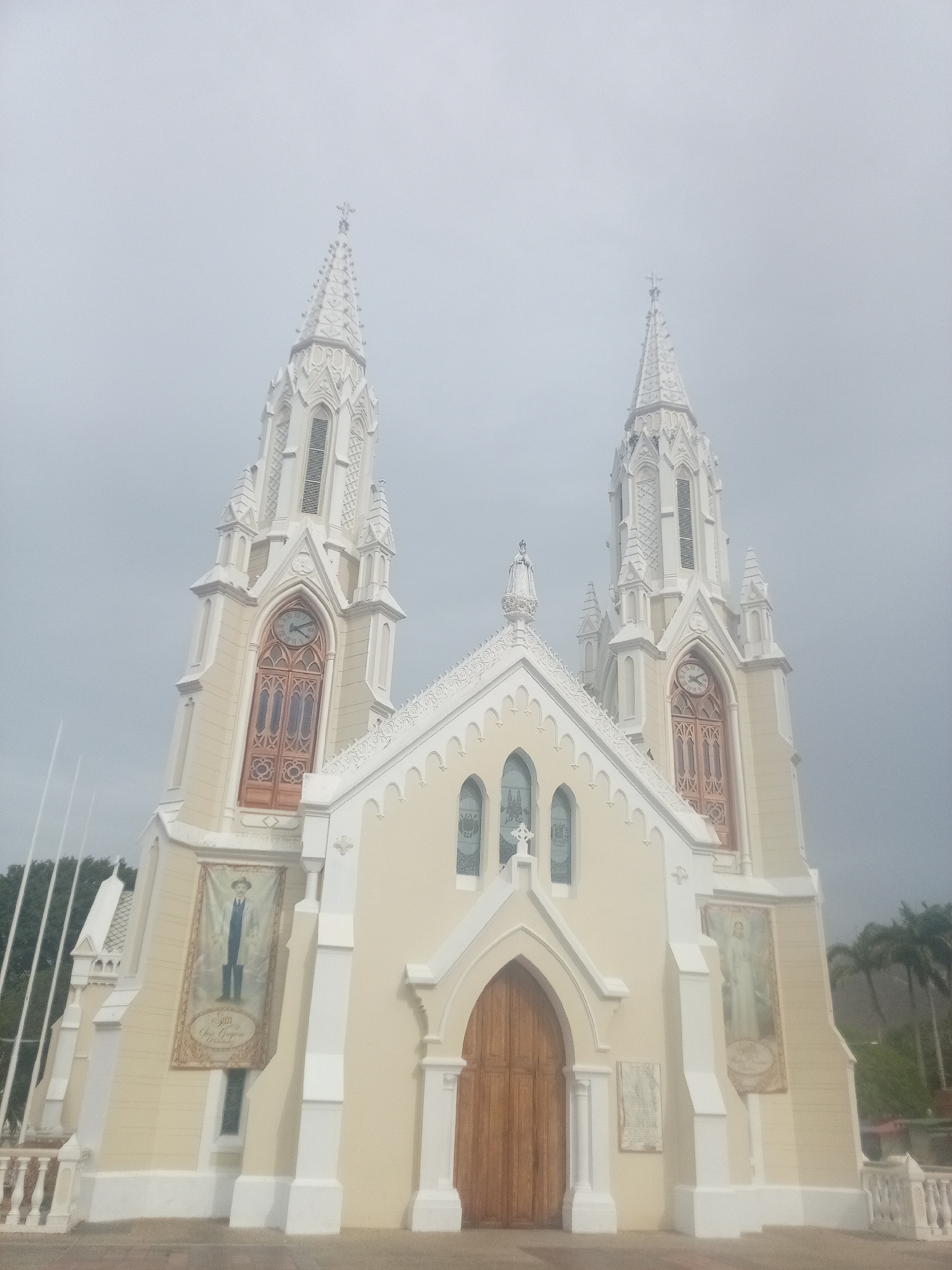 The image shows the Basilica Menor de Nuestra Señora del Valle in El Valle del Espíritu Santo, Venezuela. The basilica is a two-towered structure with Gothic Revival architecture. The building's facade is painted in light yellow with white trim. Each tower has a clock face and spires topped with crosses. Banners are hanging on either side of the main entrance. The sky is overcast. Trees can be seen in the background. A white railing surrounds the basilica.