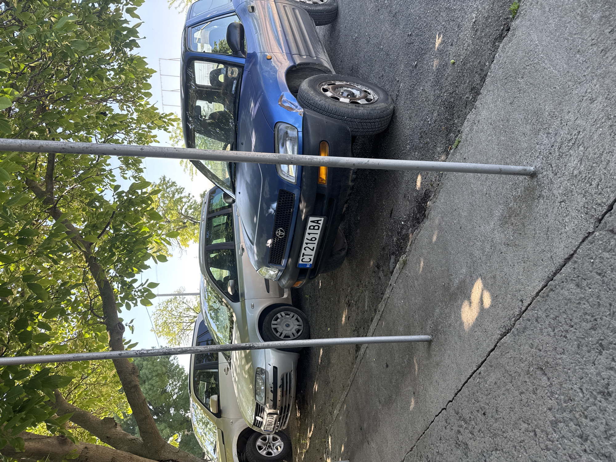 The image shows three cars parked next to each other on the side of a road. The car closest to the viewer is light-colored. Next to that is a dark-colored car and then a blue car. The license plate on the middle car reads "CT2161BA". The cars are parked next to a sidewalk and a tree. There is a metal railing in the foreground. Given that the license plate appears to be Bulgarian, it is likely that the location is indeed Sarnevo, Bulgaria.