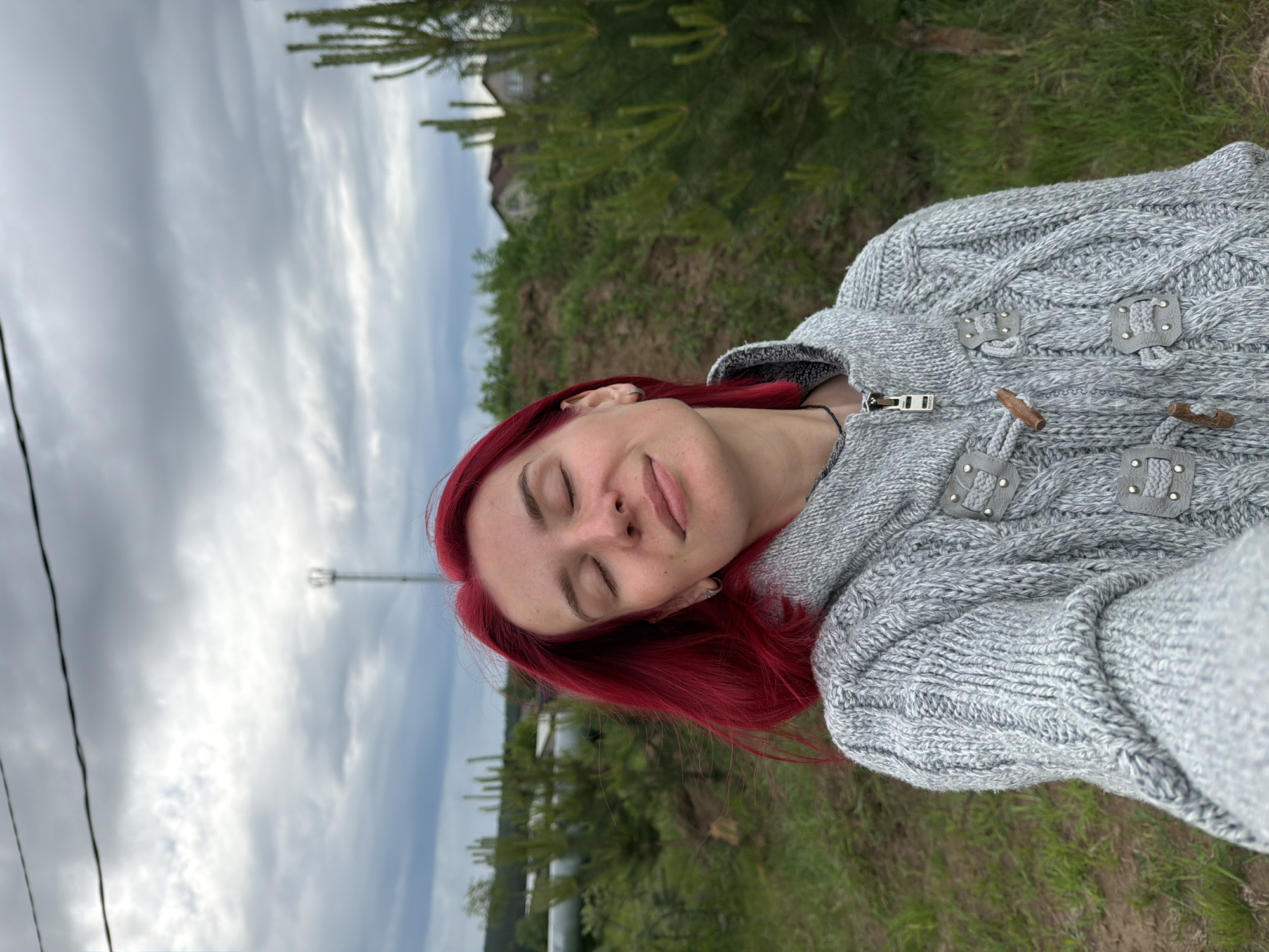 A selfie of a woman with red hair. She is wearing a grey cable knit hooded sweater with toggle closures. Her eyes are closed, and she appears to be outdoors in a grassy area. Behind her is an overcast sky, green foliage, a utility pole, and a glimpse of a building. Based on the metadata, the location is the city of Chaikovsky, Russia.
