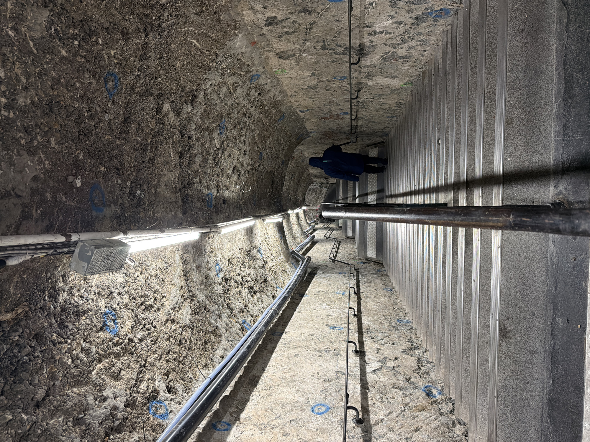 The image depicts a tunnel or underground passage, likely within Paris, France. The tunnel has a rough, rock-like wall on the left side, marked with blue circles, and a vertical, metallic wall on the right. A person in a dark coat with a hood is walking in the middle of the tunnel towards the end. A metal railing is present along the tunnel's right side, possibly for safety or guidance. Lighting fixtures are installed on the rock-like wall, illuminating the space. Pipes and other utilities run along the tunnel's left side. The floor of the tunnel appears to be a mix of concrete and exposed rock.