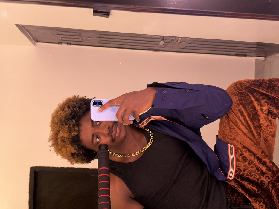 An individual, appearing to be male with dark skin and two-toned afro-textured hair (dark brown with lighter highlights), is captured in an indoor mirror selfie in Igboefon, Nigeria. He displays a neutral expression, featuring a thin mustache and soul patch. The individual wears a black sleeveless tank top, a prominent gold-colored figaro chain, and rust-brown velvet-like trousers with an embossed geometric pattern. A navy blue long-sleeved button-up shirt with striped trim (red, beige, black) is draped over his left shoulder and arm. He holds a lavender-colored smartphone with an Apple logo and dual camera lenses in his right hand, positioned to reflect the scene. In his left hand, he holds a black object with a red padded grip, consistent with a barbell or dumbbell handle. The background, visible through the mirror, consists of a plain light-colored wall and a dark panel door with vertical recesses, from which keys are hanging.