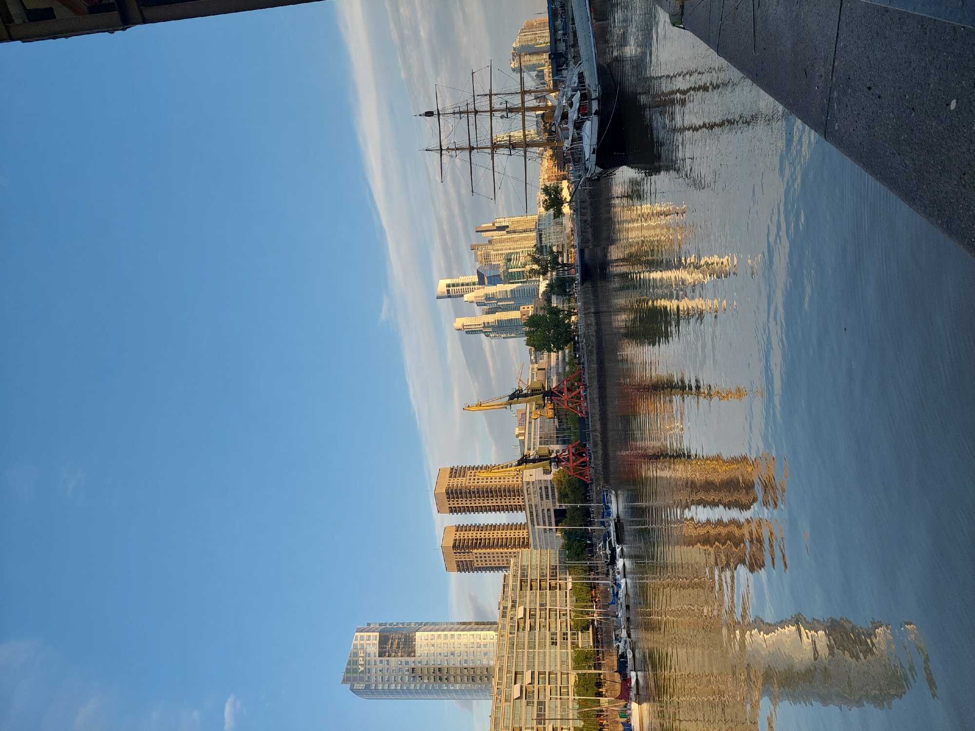 The image shows a waterfront scene in Puerto Madero, Buenos Aires, Argentina. The camera angle is slightly tilted. There are modern high-rise buildings lining the water's edge, reflected in the water. A tall sailing ship with multiple masts is docked along the pier. There are several cranes on the waterfront. The sky is blue with wispy clouds.