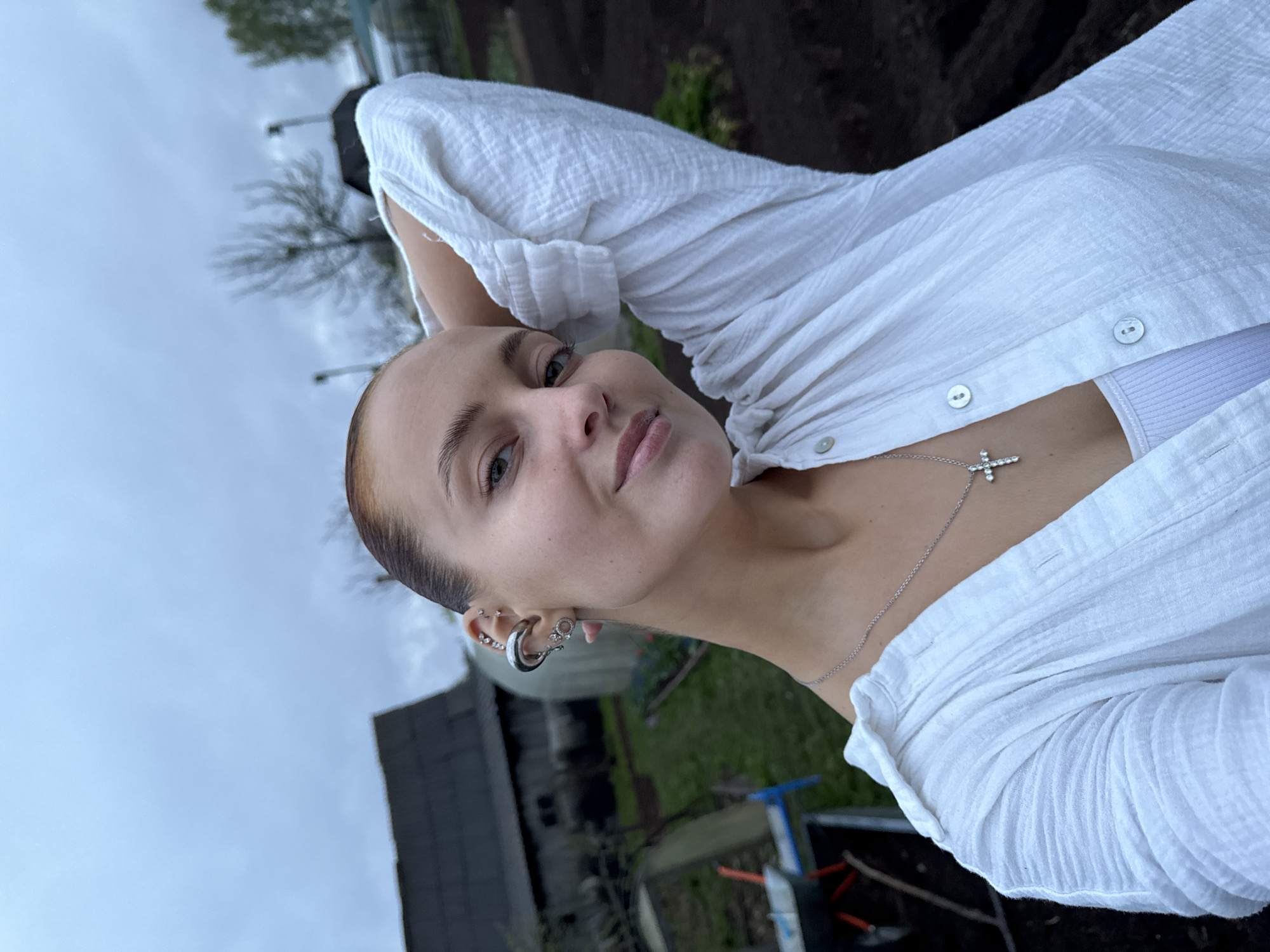 The image shows a woman outdoors. She has fair skin, brown eyes, and short brown hair slicked back. She wears a white, button-down shirt partially unbuttoned, revealing a white tank top underneath, and a silver necklace with a cross pendant. She has multiple earrings on her left ear. Her right hand is behind her head. The background includes a cloudy sky, trees, and what appears to be a garden area with dark soil and a structure possibly related to gardening. The location is potentially Chastyye, Russia.