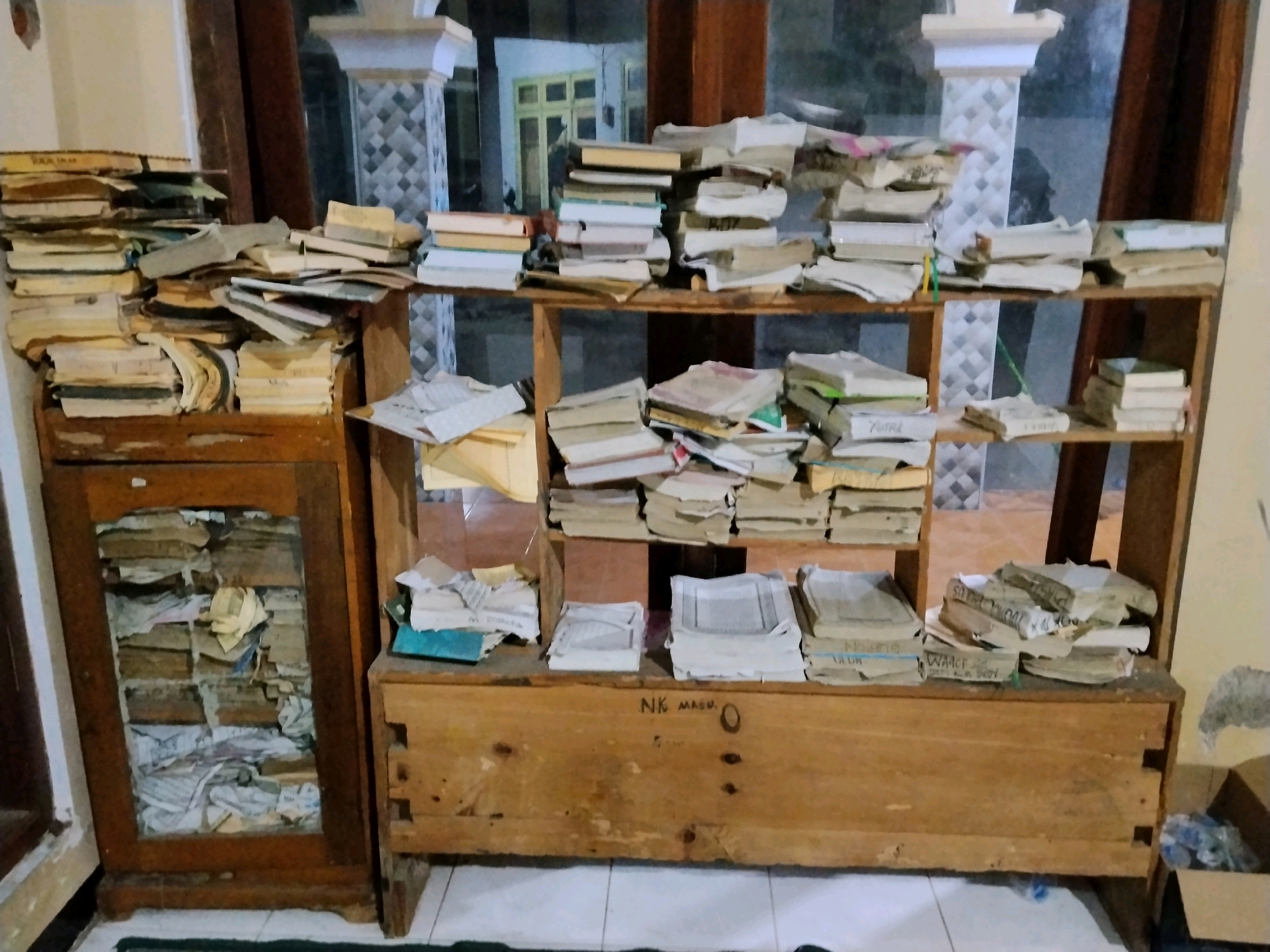 The image depicts a cluttered room with a significant amount of books and papers organized on wooden shelves and within a wooden cabinet. The room appears to be an indoor space, possibly a study or a storage area.
On the left side of the image, a wooden cabinet with a glass door displays stacks of papers and possibly books inside. The cabinet has a dark wooden frame and is situated on the floor.
To the right of the cabinet, there are several wooden shelves filled with books and loose papers. The books are stacked in various orientations, some upright, some lying flat, and many are in disarray. The papers are also piled haphazardly.
In the foreground, a long, low wooden chest or desk is visible. The words "NK MASU" are written on its surface. Several stacks of papers are placed on top of this chest.
The background of the image shows what appears to be a window or doorway with a view of another room or area. Architectural elements like pillars and patterned tiles are visible in the background, suggesting an indoor setting with some decorative features. The lighting in the room is dim, creating shadows and highlighting the textures of the wood and paper.
The overall impression is one of disorganization and a large collection of reading material. There are no people or animals visible in the image. The presence of the text "NK MASU" on the wooden chest might be a label or inscription. The scene suggests a place where information is stored or studied, but it is currently in a state of considerable clutter.