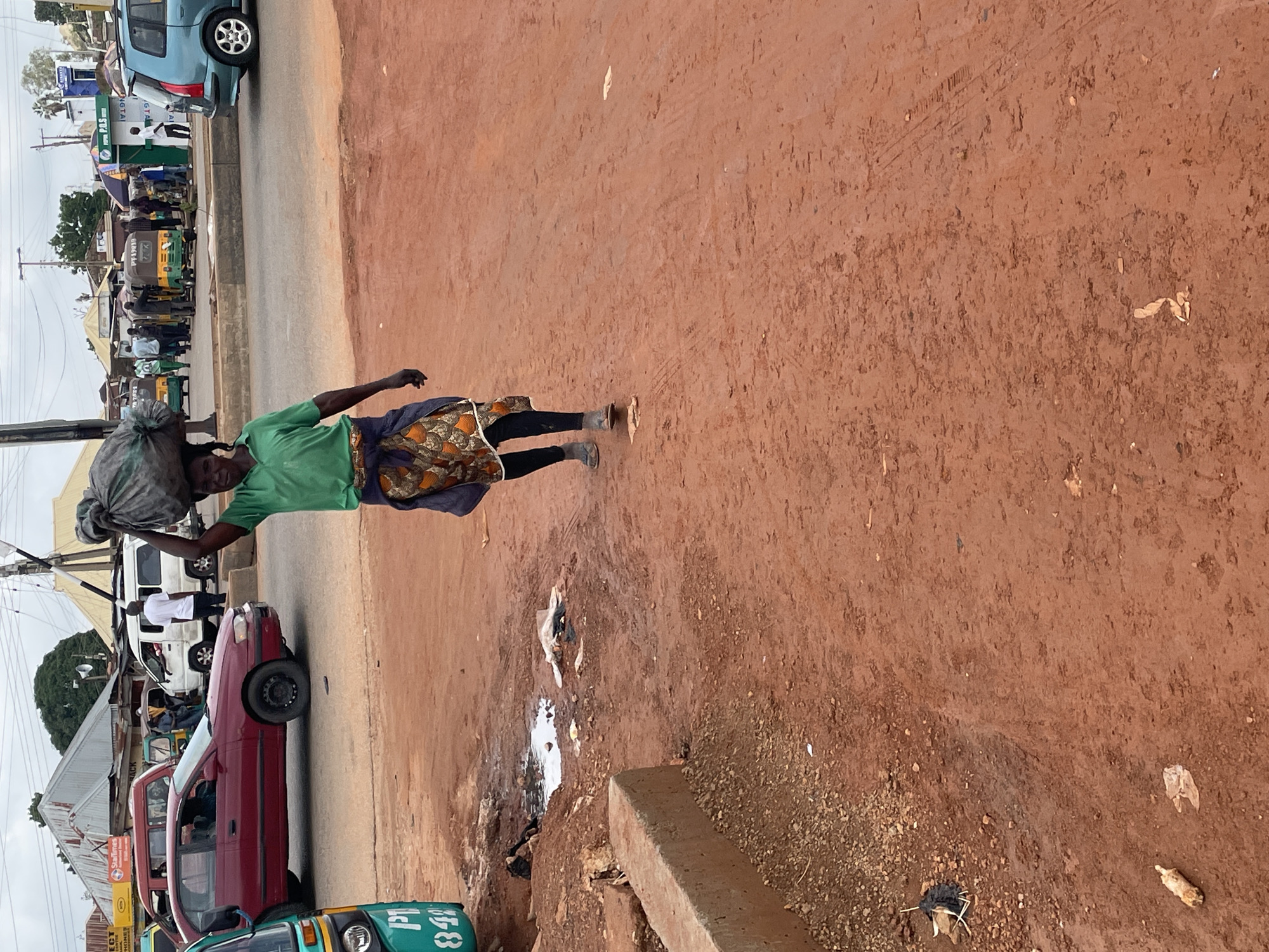 A woman is walking on a dirt road in Gura Top, Nigeria, carrying a large bag of charcoal on her head. She is wearing a green shirt and a patterned skirt. In the background, there are several vehicles, including a red car, a blue SUV, and a few auto-rickshaws. There are also some buildings and people standing around. The sky is cloudy. The ground is made of red dirt and there are some puddles of water.