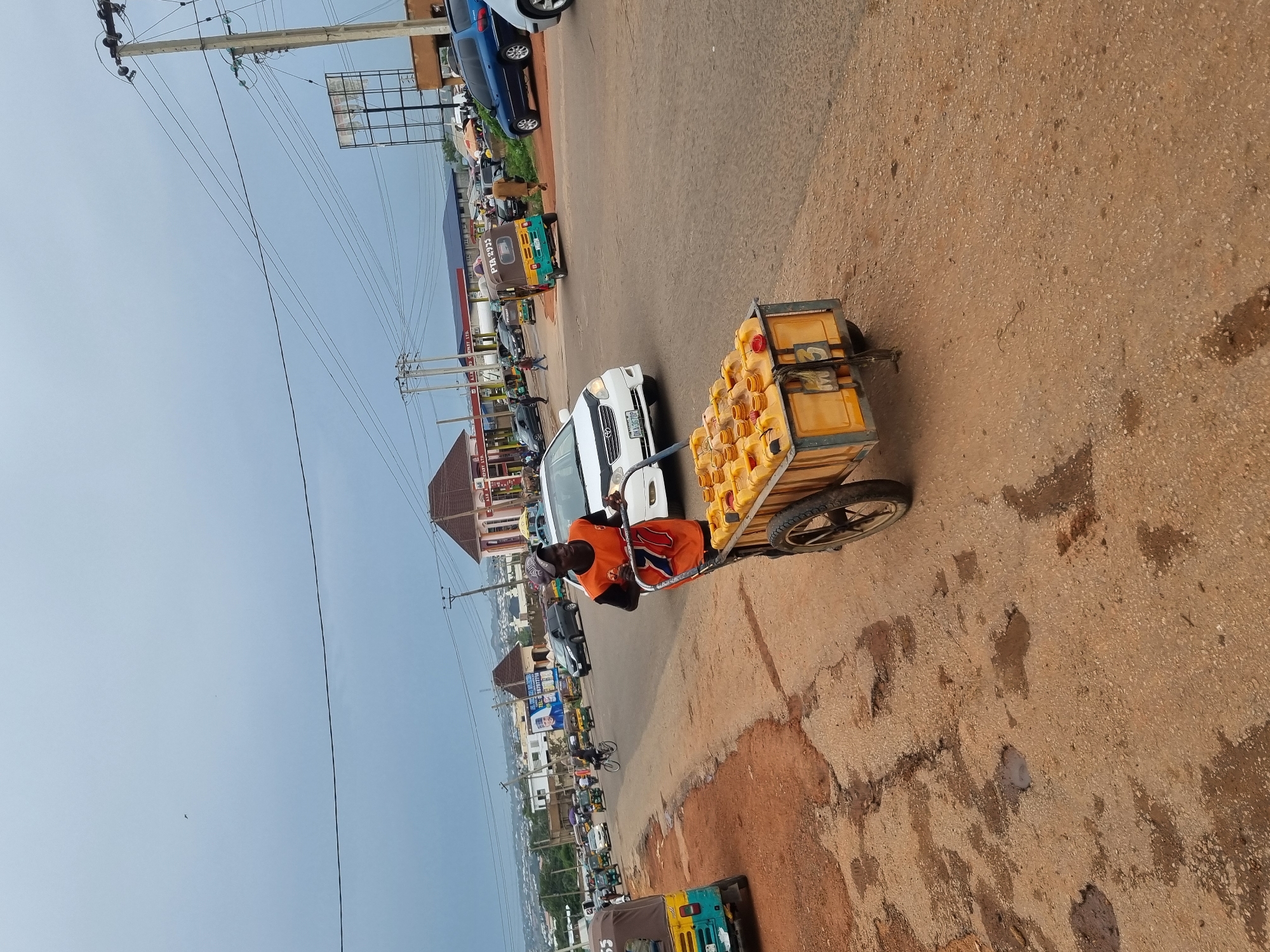 In Jos, Nigeria, a man pushes a cart laden with yellow jerry cans of fuel down a dusty road. The cart has two large wheels and a metal frame. The man, wearing an orange shirt and a cap, is walking behind the cart. Several vehicles, including cars and a tricycle, are visible on the road in the background. Buildings and utility poles line the street. The sky is overcast, suggesting a cloudy day. The road surface is uneven and appears to be a mix of dirt and gravel, with some puddles visible, indicating recent rainfall. The scene depicts a common mode of commerce and transportation in the region.