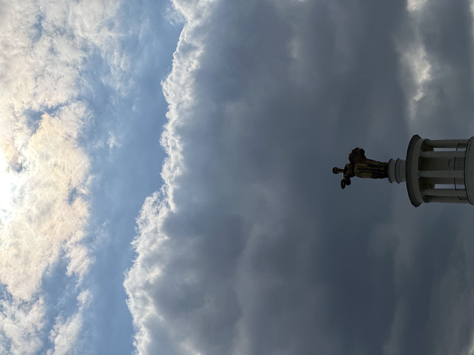 The image is a tilted, low-angle shot of the Pennsylvania State Capitol in Harrisburg, Pennsylvania. The golden statue of Commonwealth is visible atop the capitol dome, silhouetted against a backdrop of dramatic clouds. The sky transitions from a bright, sunlit area with scattered clouds on the left to a much darker, ominous storm cloud formation on the right. The architecture of the dome, including its columns and detailed ornamentation, is partially visible.