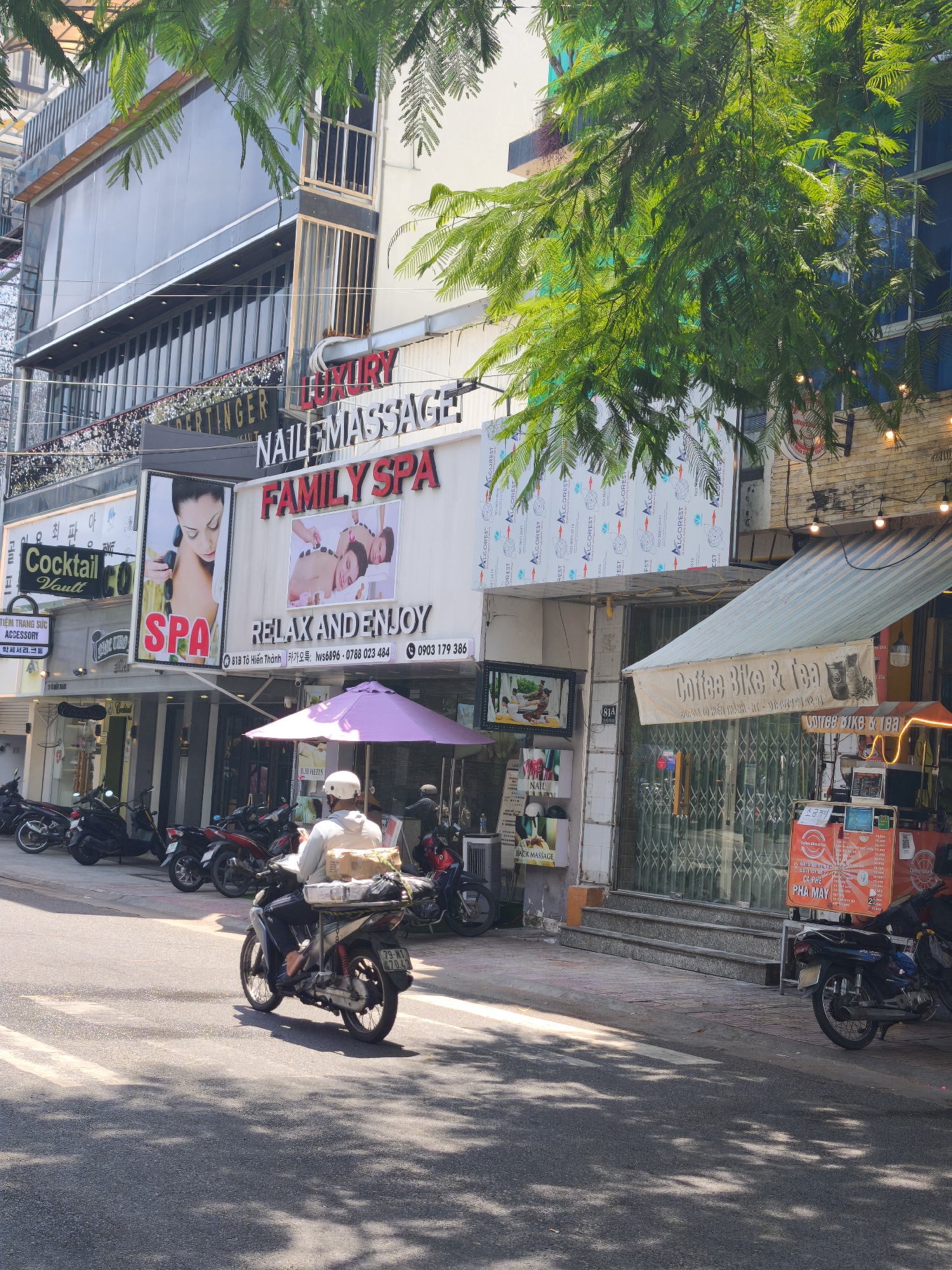 The image shows a street scene in Nha Trang Ward, Vietnam, on a bright, sunny day, evidenced by the strong shadows cast on the road and pavement. The primary focus is on the storefronts lining the street, with a prominent "LUXURY NAILEMASSAGE FAMILY SPA" sign dominating the upper portion of the scene. Adjacent to this is a "Cocktail Vault" sign. Below the spa sign, "RELAX AND ENJOY" is advertised, along with an address and phone numbers in Vietnamese and Korean.
The street is lined with parked motorcycles, indicating a common mode of transportation. A person riding a motorcycle, carrying packages on the back, is moving away from the camera down the street, contributing to the sense of daily life in the area.
Further down the street, another establishment with a sign reading "Coffee Bike & Tea" is visible, featuring a striped awning and outdoor seating. The windows of the shops are largely covered by metal grilles, a common security feature. The overall impression is a bustling commercial street in a Vietnamese city, with a mix of businesses catering to local needs and possibly tourists, given the presence of Korean text. The lighting suggests it is daytime, likely mid-morning or afternoon.