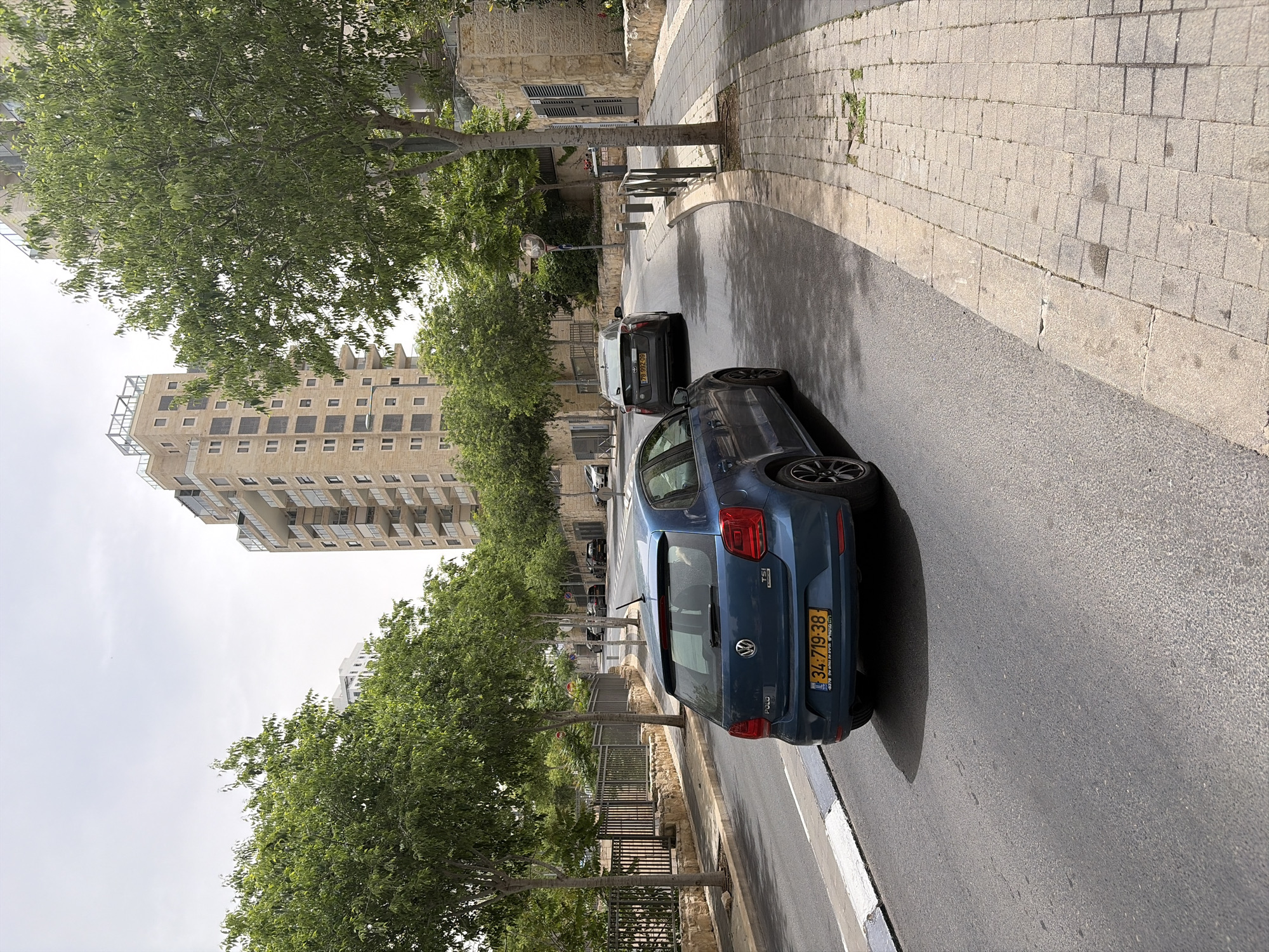 The image depicts a street scene, likely in an urban setting. A blue Volkswagen Polo is parked on the right side of the road. Behind it is a black car. Tall buildings are visible in the background, alongside lush green trees lining the street. The road is asphalt, and the sidewalks are paved with rectangular blocks. The license plate on the Polo reads "34-719-38".