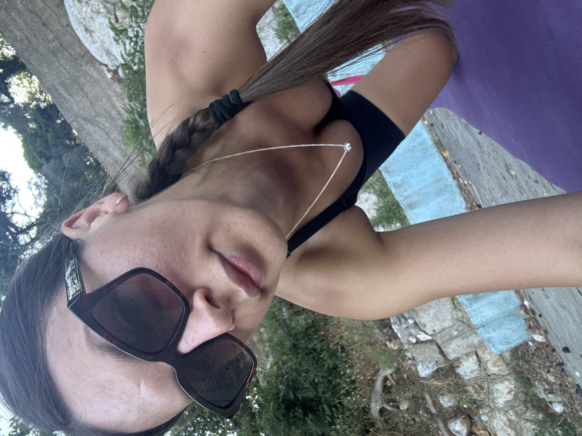 A woman with long dark hair in a braid, wearing sunglasses and a black sports bra, kneels on a purple yoga mat outdoors. She is positioned at an angle, with her face and upper torso in the foreground. The background shows greenery, rocks, and a stone wall. The setting appears to be a natural or semi-natural area, possibly a park or hillside, in Boreti, Montenegro.