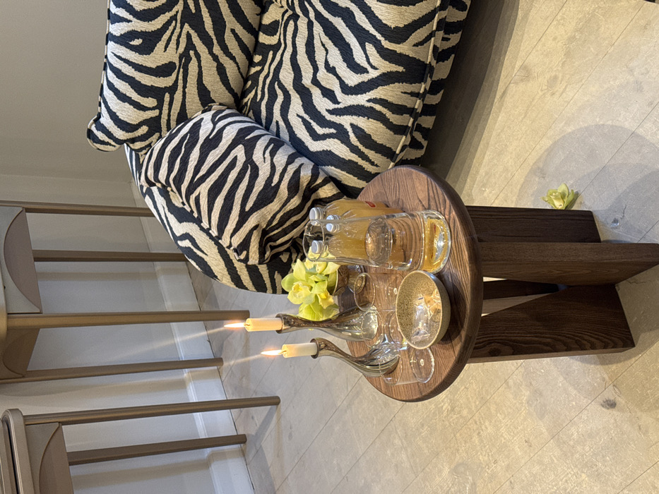 This image depicts an indoor scene in the city of Unknown, Unknown, featuring modern furniture and refreshments. In the foreground, a round, dark brown wooden side table with three angled legs is visible. Positioned on the table are two lit white taper candles in distinct silver-colored candleholders, a small metallic bowl containing yellow-green flora, two clear empty drinking glasses, a small speckled beige bowl holding wrapped items, one large clear glass bottle containing clear liquid, and two smaller clear glass bottles containing an orange-yellow liquid.
To the right of the side table, a large upholstered sofa or armchair with a black and beige zebra-stripe pattern covers the majority of its visible surface and cushions. In the background, against a light-colored wall, two tall, narrow console tables with light-toned frames and curved front panels are present. The floor is a light grey, smooth material. A single yellow-green petal or flower fragment rests on the floor next to the base of the side table. No individuals are present in the scene.