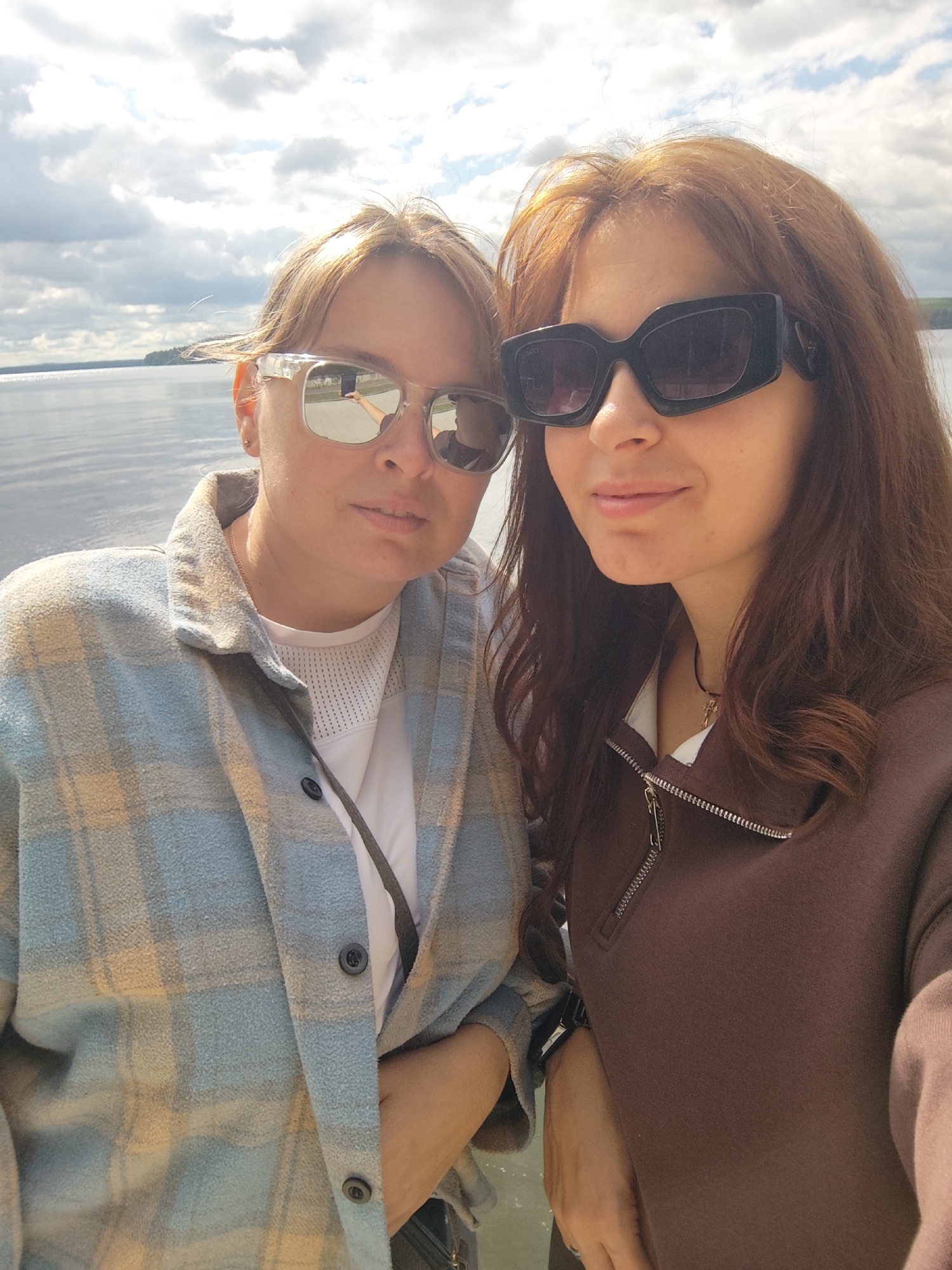 Two women are taking a selfie outdoors. The woman on the left has blonde hair and is wearing sunglasses, a blue and yellow plaid shirt, and a white shirt underneath. The woman on the right has brown hair and is wearing large black sunglasses and a brown zippered jacket over a white shirt. They are standing in front of a body of water with a cloudy sky in the background. The location is likely Chaikovsky, Russia, given the context of other images from the same source.