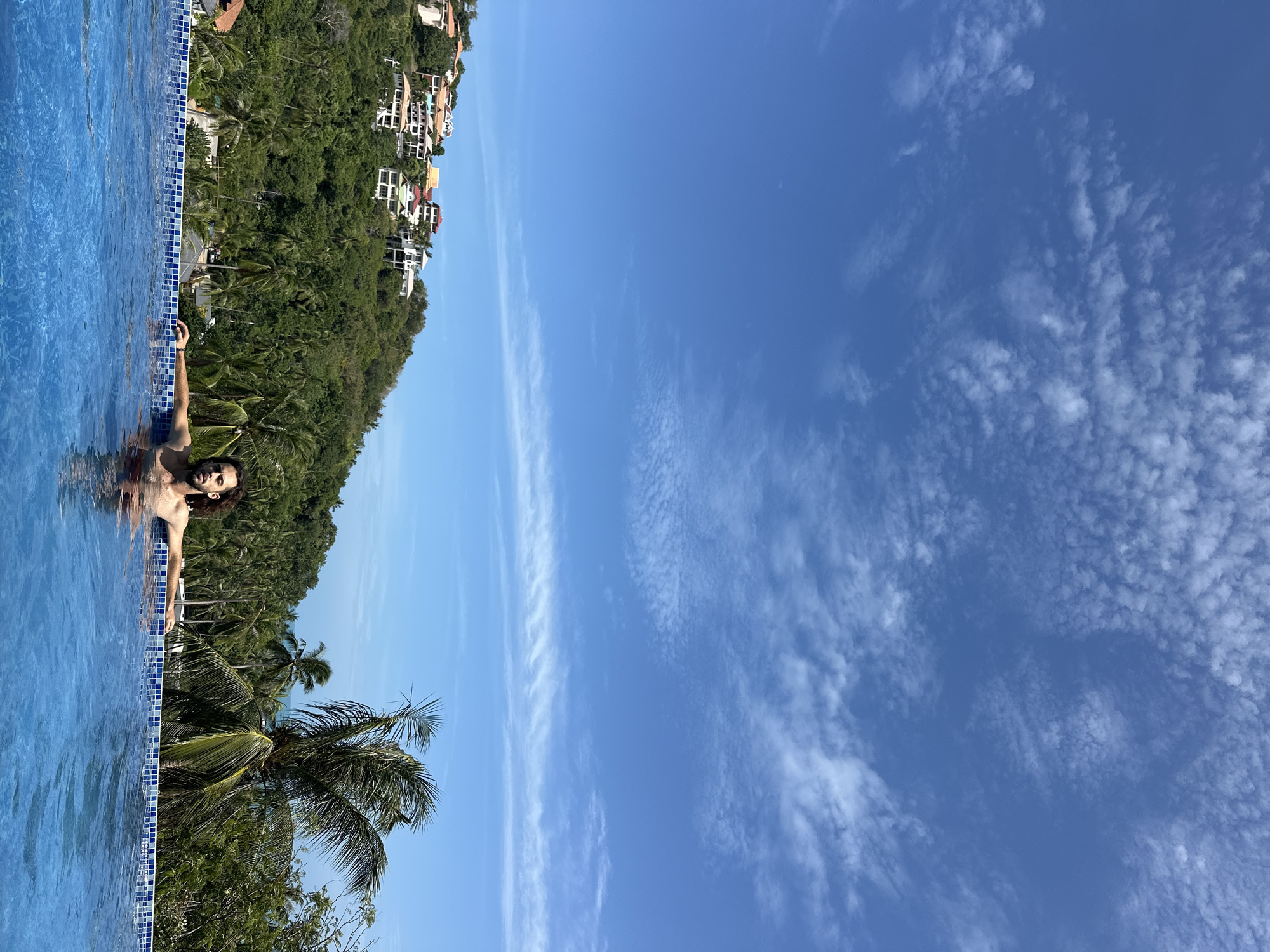 A man with dark, curly hair is partially submerged in a bright blue infinity pool, his arms resting on the tiled edge. He is looking directly at the camera, his expression neutral. Behind him, a lush green hillside is dotted with white and red-roofed houses, suggesting a residential area. Palm trees are scattered throughout the landscape, and the vast blue sky above is streaked with thin, wispy white clouds. The scene appears to be a resort or private residence overlooking a tropical landscape, likely in the Ko Samui District of Thailand.