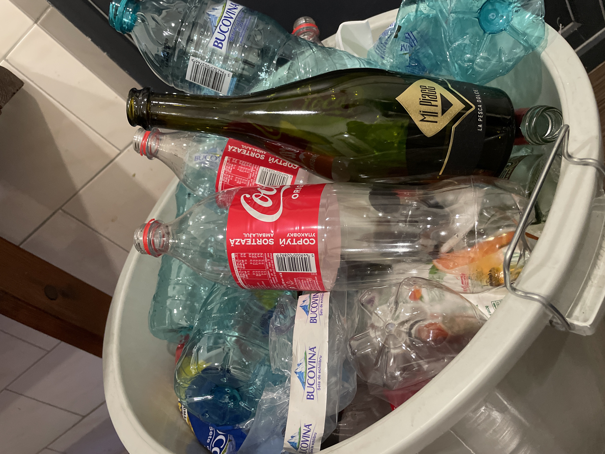 A close-up, overhead shot shows a white plastic trash bin filled with various empty plastic and glass bottles in Vadul lui Voda, Moldova. Among the bottles are several clear plastic water bottles, one with a blue cap and a "BUCOVINA" label, and another clear plastic bottle with a red label that reads "COCA-COLA". There is also a green glass bottle with a gold label that says "Mi Piace LA PESCA DOLCE". The bottles are cluttered together, with some crushed and others still intact. The bin is made of white plastic and has a metal handle on the right side. The background shows a tiled wall and a wooden beam.