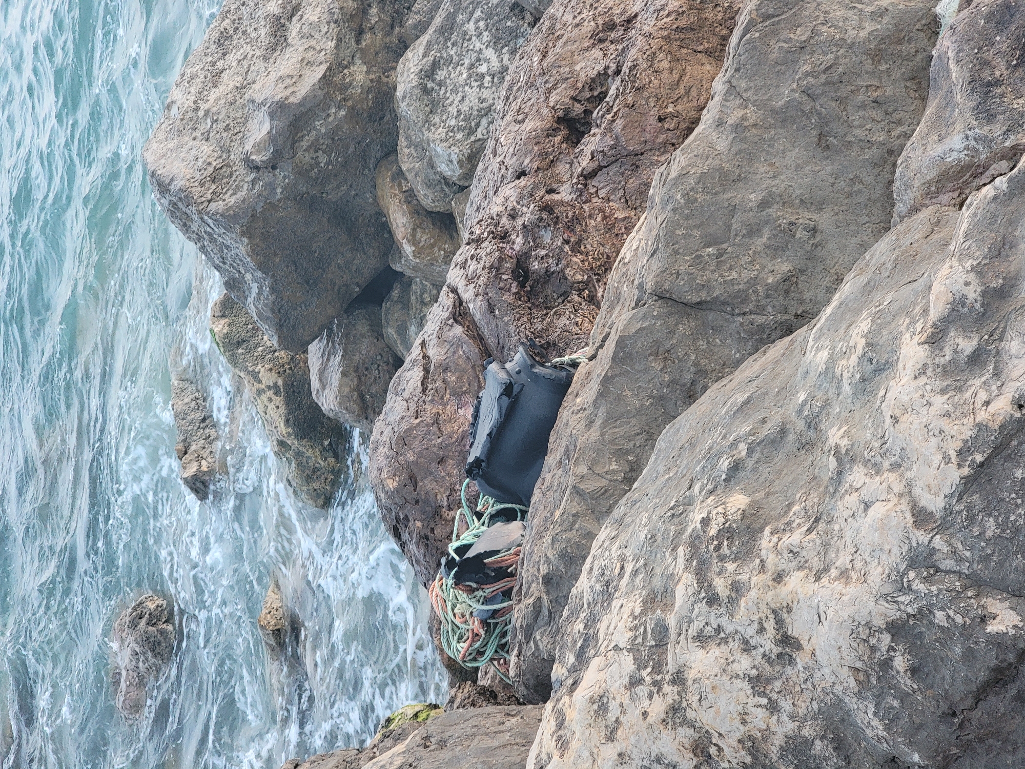 The image depicts a rocky shoreline with the sea in the background. Several large, gray rocks are scattered across the foreground and midground, some of which are partially submerged in the water. In the midground, a tangled coil of multi-colored rope, predominantly green and orange, rests on top of a dark-colored object, possibly a bag or a piece of equipment. The water is a mix of blue and white, indicating waves and currents. The overall scene is one of a natural coastal environment with some man-made debris present. There are no people visible in the image.