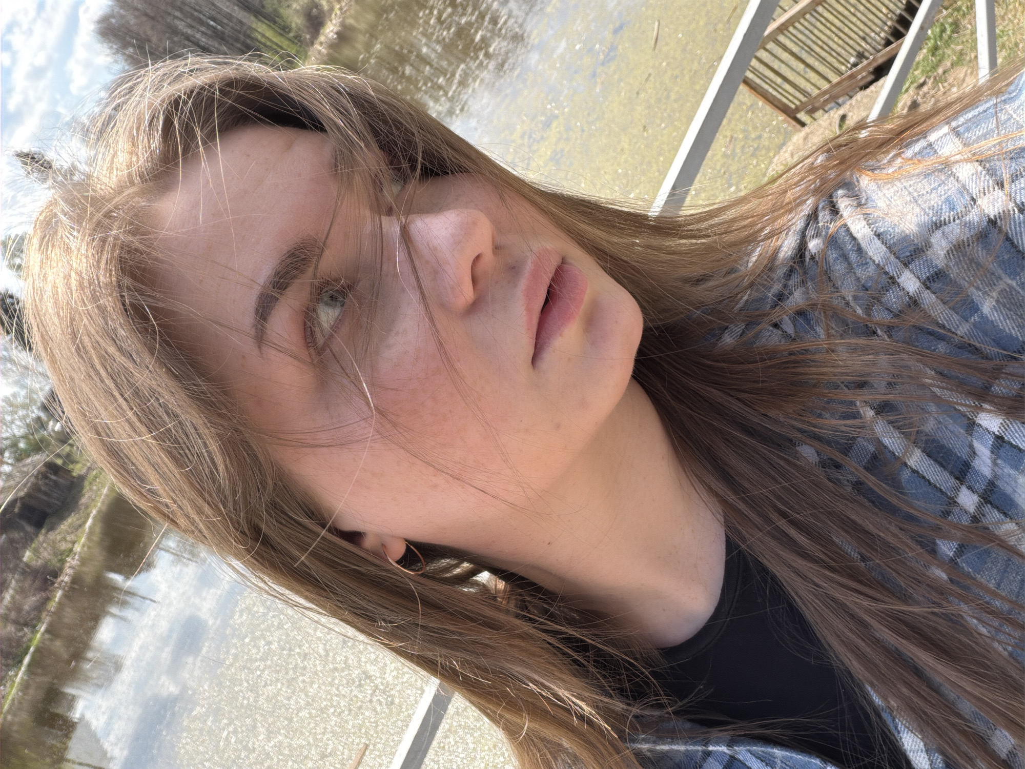 A selfie captures a young woman with light brown hair, a fair complexion speckled with freckles, and light-colored eyes. She is wearing a blue and white plaid shirt over a black top. A thin, circular earring is visible in her left ear. The background shows a body of water reflecting the sky and surrounding trees, and a metal railing is in the upper right. The location is likely in Chaikovsky, Russia, given the prompt's instructions. The woman's face is the primary subject, with the background suggesting an outdoor setting, possibly a park or lakeside area. The selfie angle indicates the woman is holding the camera.