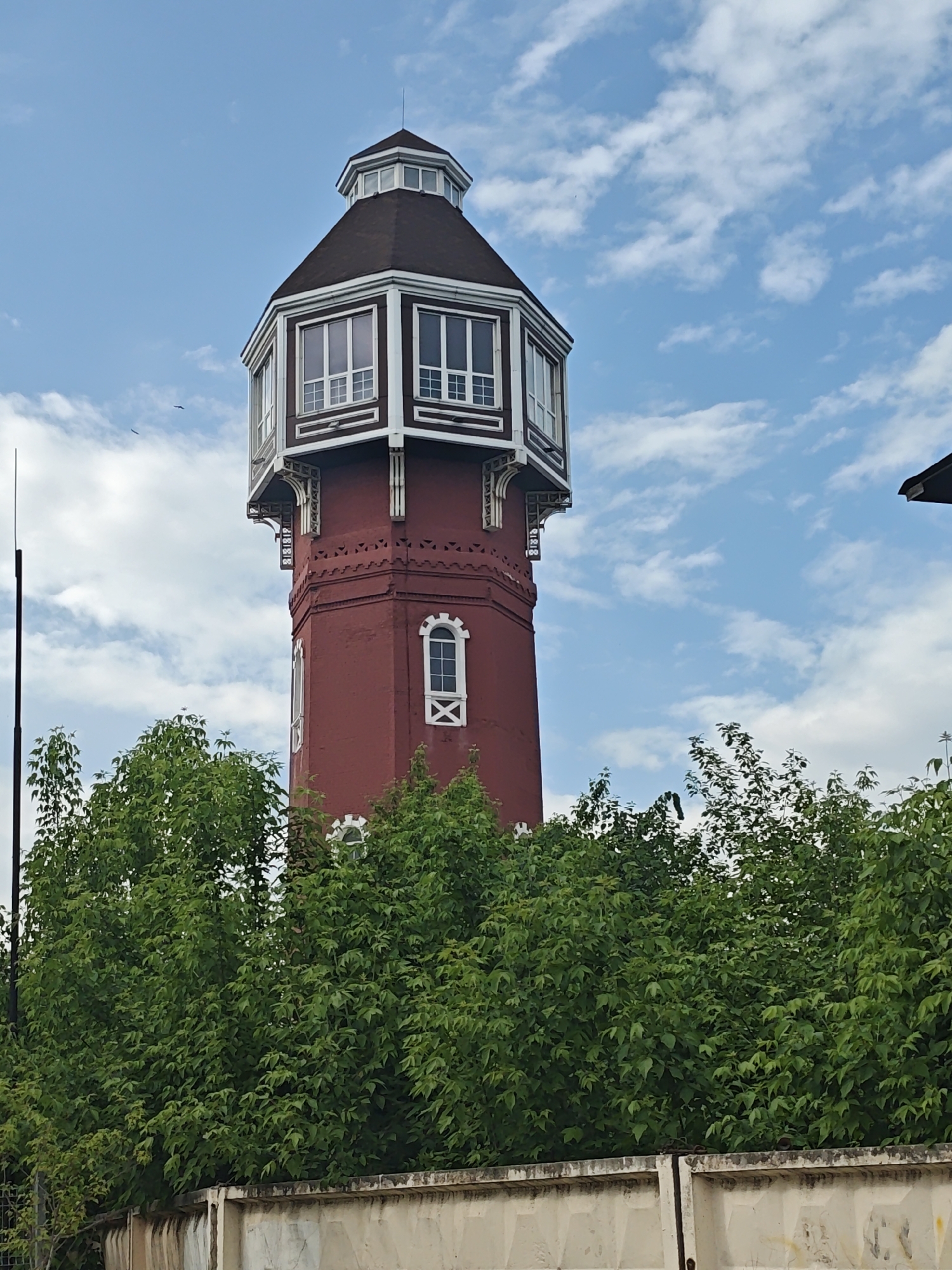 The image shows the Noginsk water tower, a historic landmark in the city of Noginsk, Russia. The tower is constructed of red brick, with a distinctive polygonal observation deck topped with a brown roof. White-framed windows are visible in the observation deck and on the tower's main structure. Lush green trees partially obscure the base of the tower, and a concrete fence runs along the bottom edge of the frame. The sky is blue with scattered white clouds. A lightning rod is attached to the side of the tower.