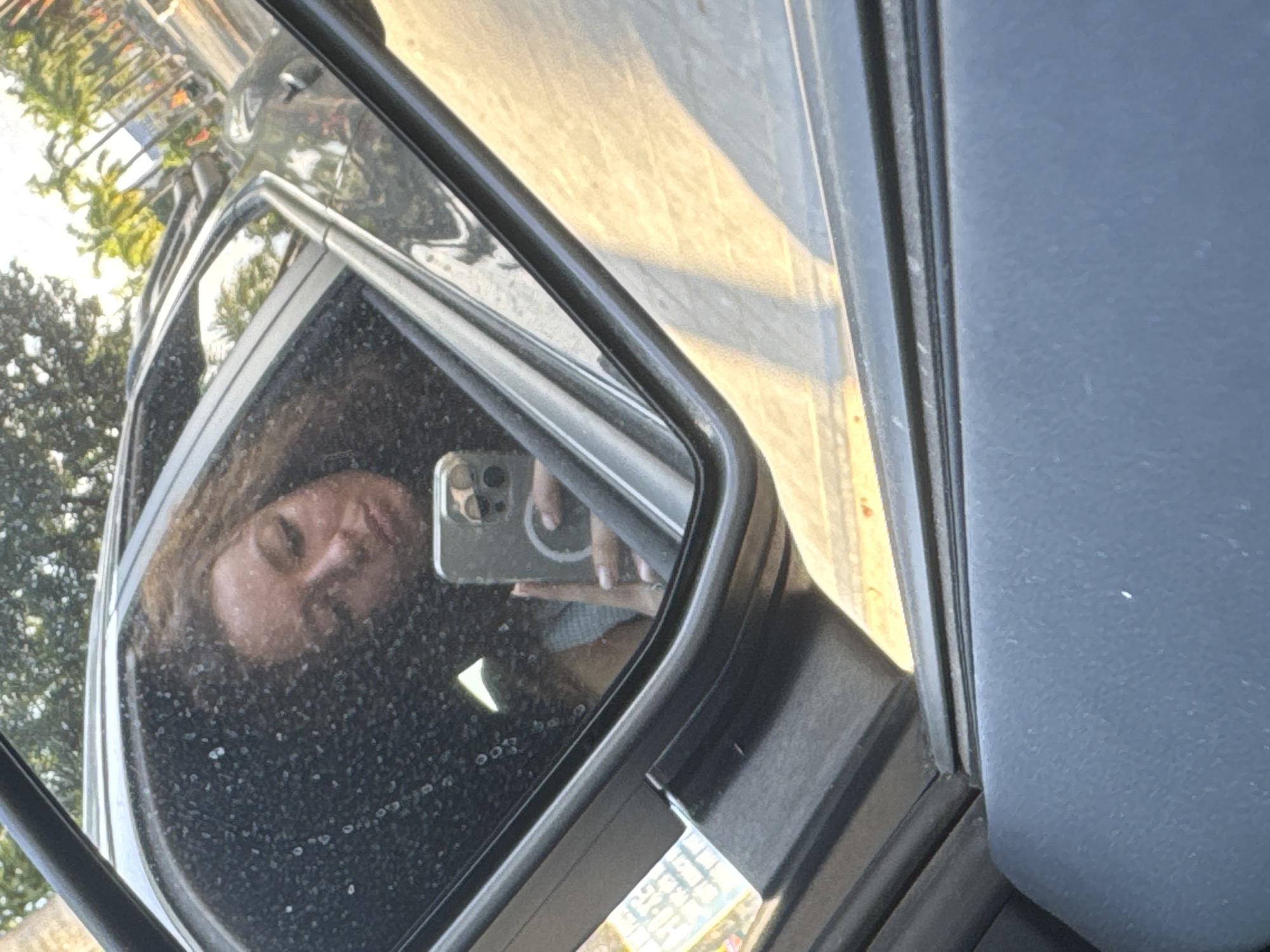 The image shows a woman reflected in a car side mirror. She is holding a smartphone, presumably taking a selfie. The background visible in the mirror suggests an outdoor urban environment with trees and buildings. A street or road can also be seen in the car's mirror. The location is approximated in the city of Lao Cai Ward, Vietnam, but without further evidence to provide confirmation or further details on the environment, this is an approximation.