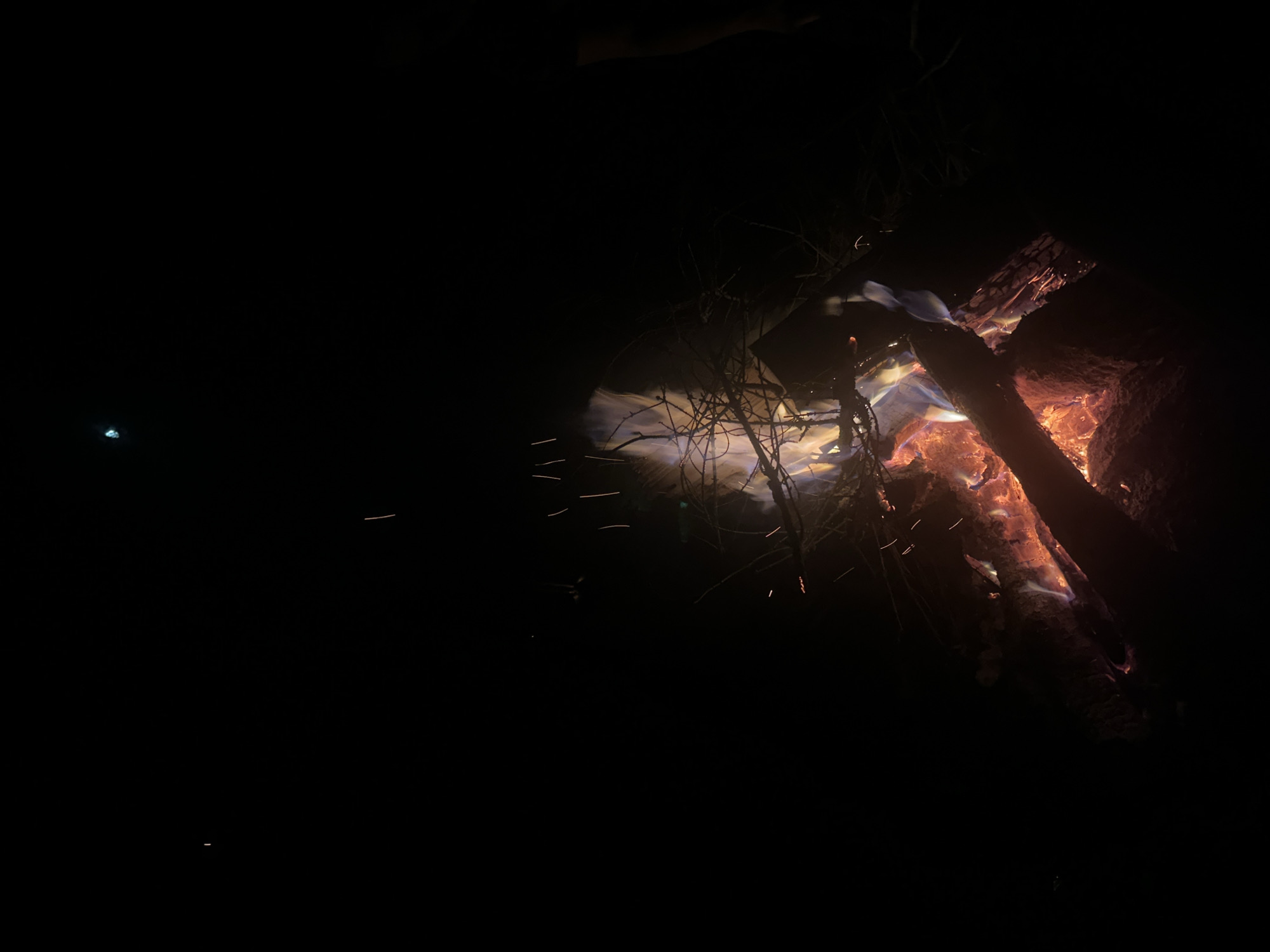 The image depicts a nighttime campfire in Юзуфоўскі сельскі Савет, Belarus. The fire is composed of burning logs and twigs, emitting orange and blue flames, along with sparks. The surrounding environment is dark, with some tree branches silhouetted against the firelight.