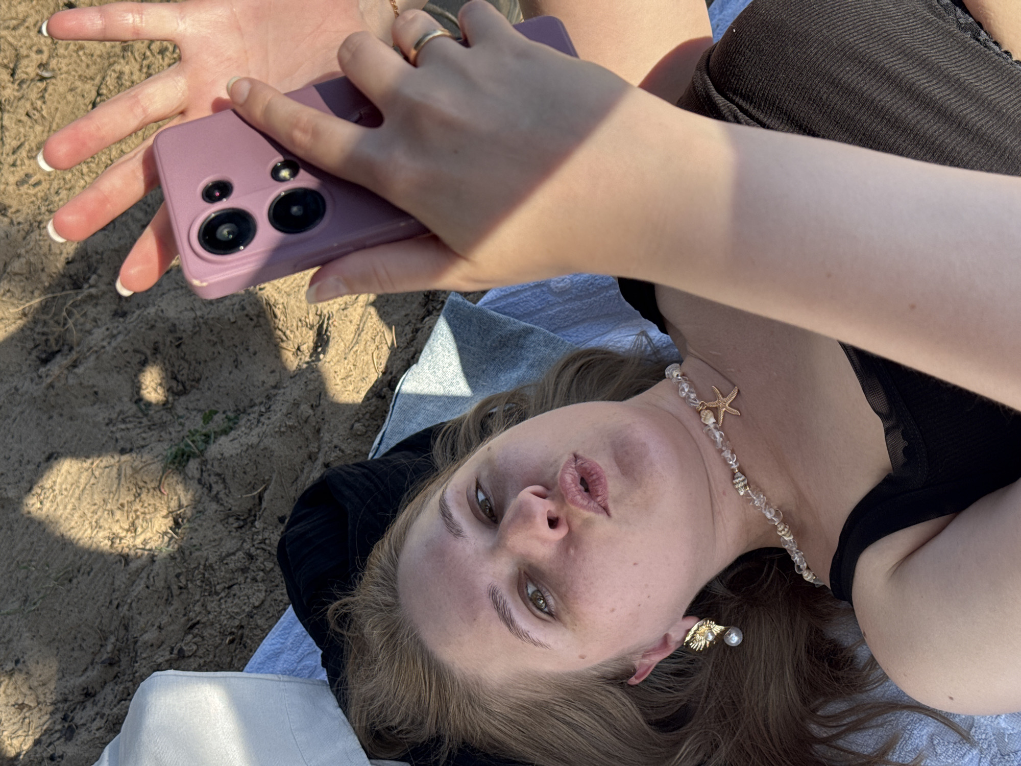 A person is lying on sand, taking a selfie with a purple smartphone. The person has blonde hair, is wearing a black tank top and a beaded necklace with a starfish pendant. There's a large white pillow on the left side of the frame and a light blue towel. Sunlight casts shadows across the sand.