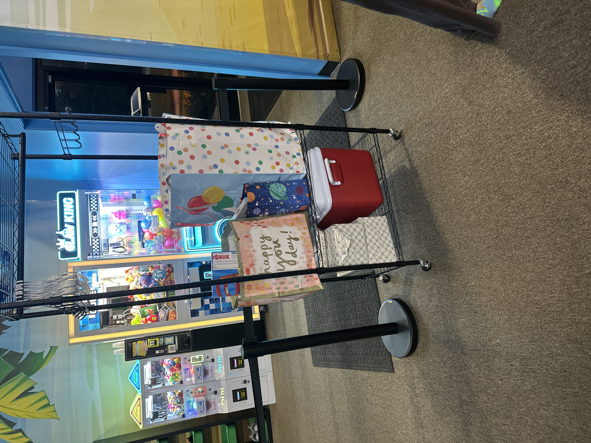 A dark grey, wheeled shelving unit is positioned indoors on a textured, grey carpet. The unit holds several gift bags, a red cooler, and a white gift box. Behind the shelving unit, several arcade machines with bright lights and colorful prizes are visible. A black stanchion with a retractable belt is in the foreground, to the left of the shelving unit. The background includes a wall with a mural and a doorway. The scene appears to be in a commercial establishment, potentially an arcade or entertainment venue in Buffalo Grove, United States of America.