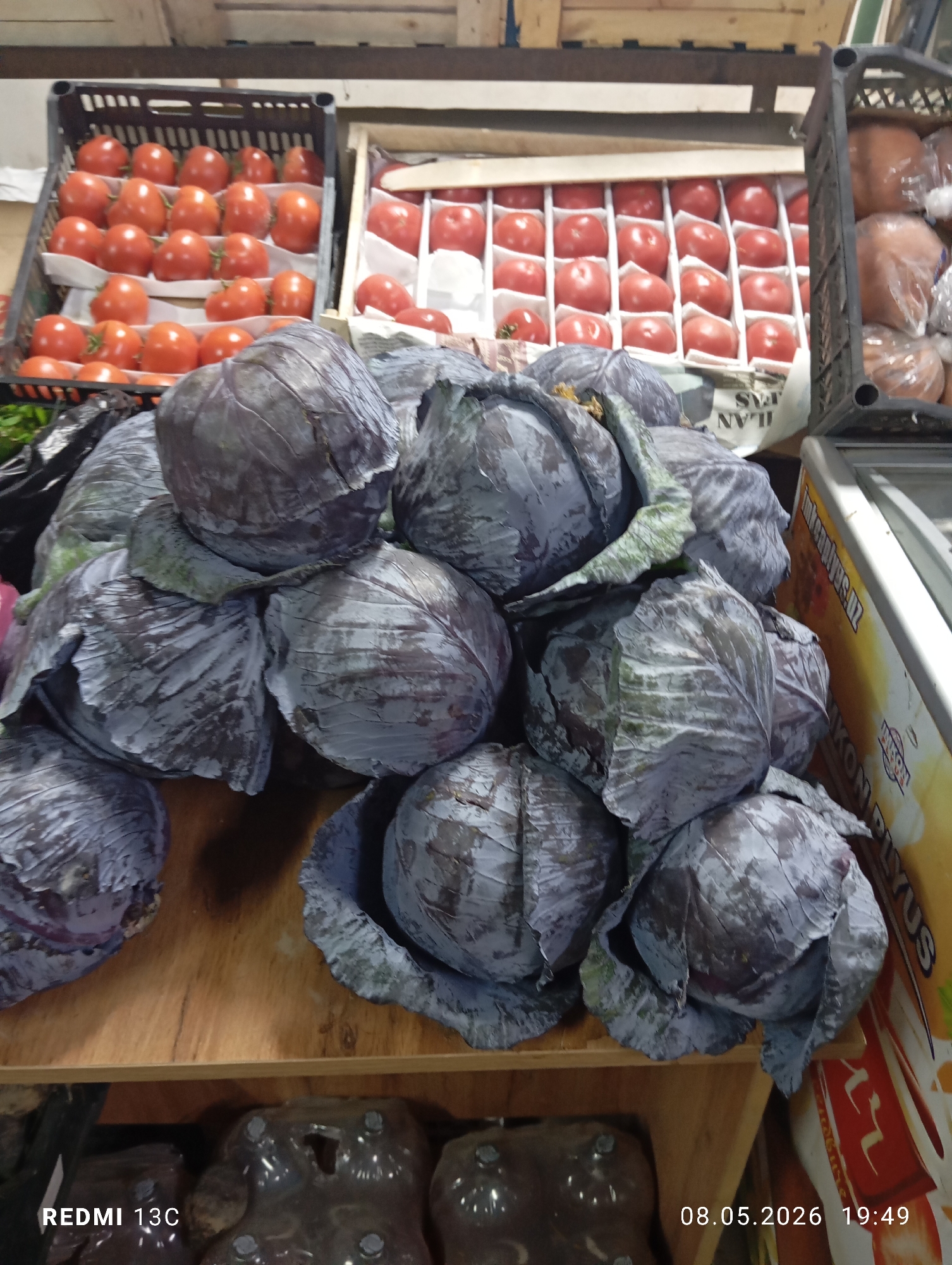 The image shows a display of red cabbages in what appears to be a market setting, based on the presence of other produce like tomatoes. The cabbages are piled on top of each other on a wooden surface. Additional merchandise is visible in the background, including tomatoes arranged in crates or boxes. The image was captured in Gazalkent, Uzbekistan, as indicated by the writing on the side of the refrigerator. The photo was taken on 08.05.2026 at 19:49.