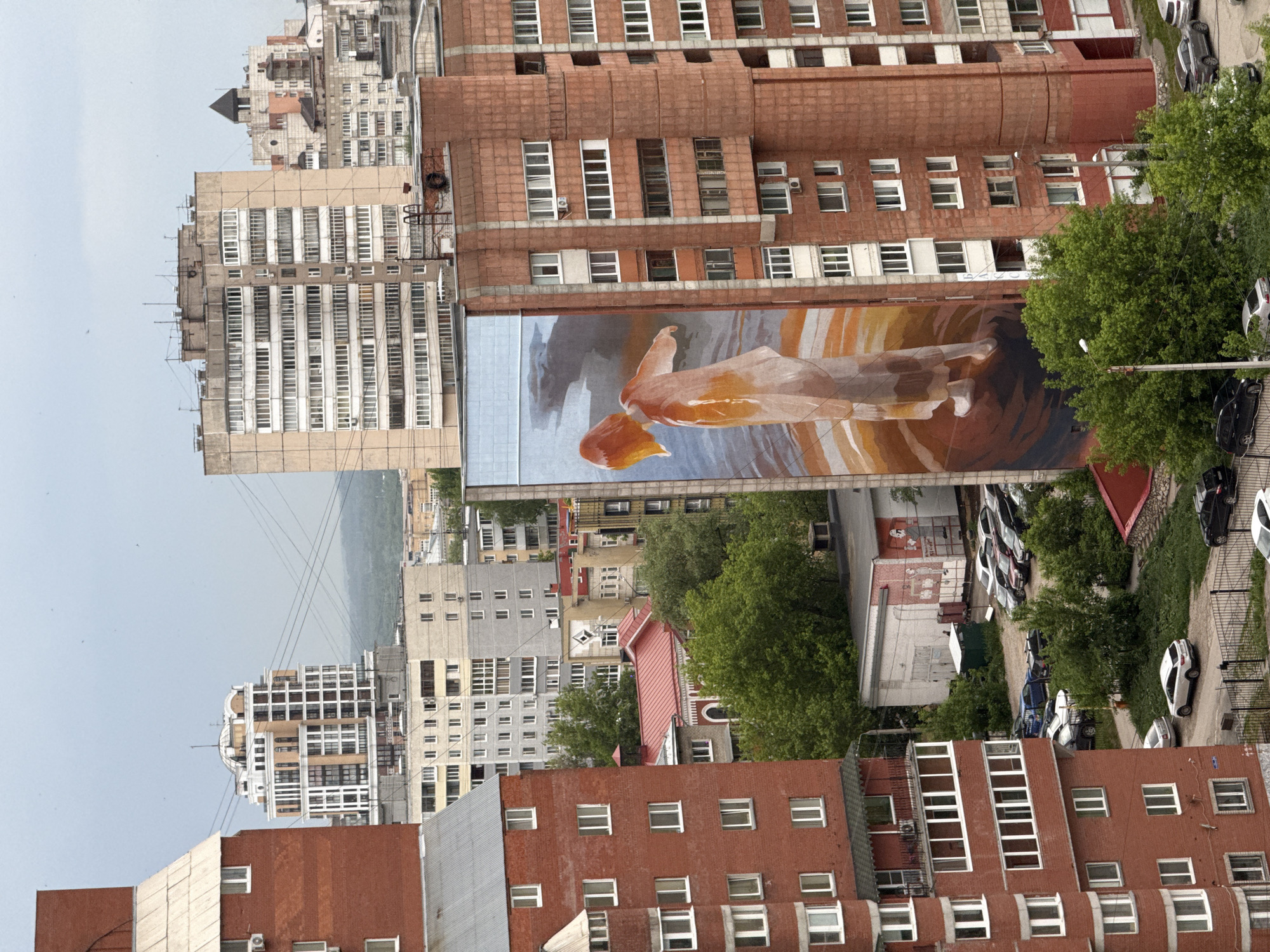 This is an exterior, high-angle shot of a city scene, with numerous buildings, streets, and vehicles. A large mural is prominently featured on the side of a tall, brick-colored building. The mural depicts a figure in flowing, orange and brown tones, set against a blue and brown background. A street with parked cars runs adjacent to the mural, with trees lining the street. In the background, a mix of buildings of varying heights and architectural styles fill the urban landscape, set against a muted sky. This photograph was taken in the city of Perm, Russia.