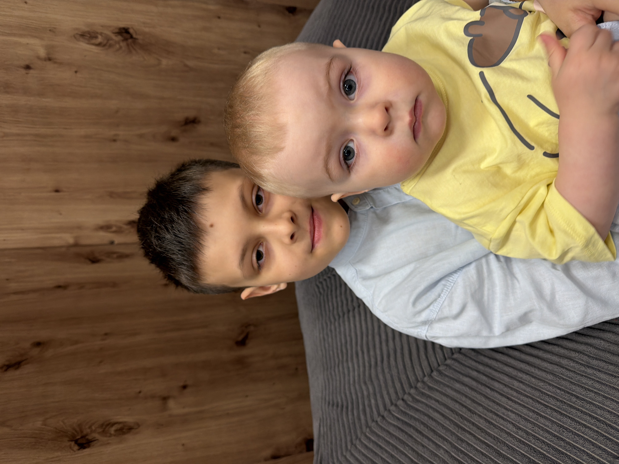 Two children are positioned together on a textured grey surface. The younger child, with blonde hair and wearing a yellow t-shirt, is angled slightly towards the viewer. The older child has dark hair and wears a light blue shirt. The background is a wooden surface. The location is unspecified and there is no indication of Votkinsk, Russia in the image.