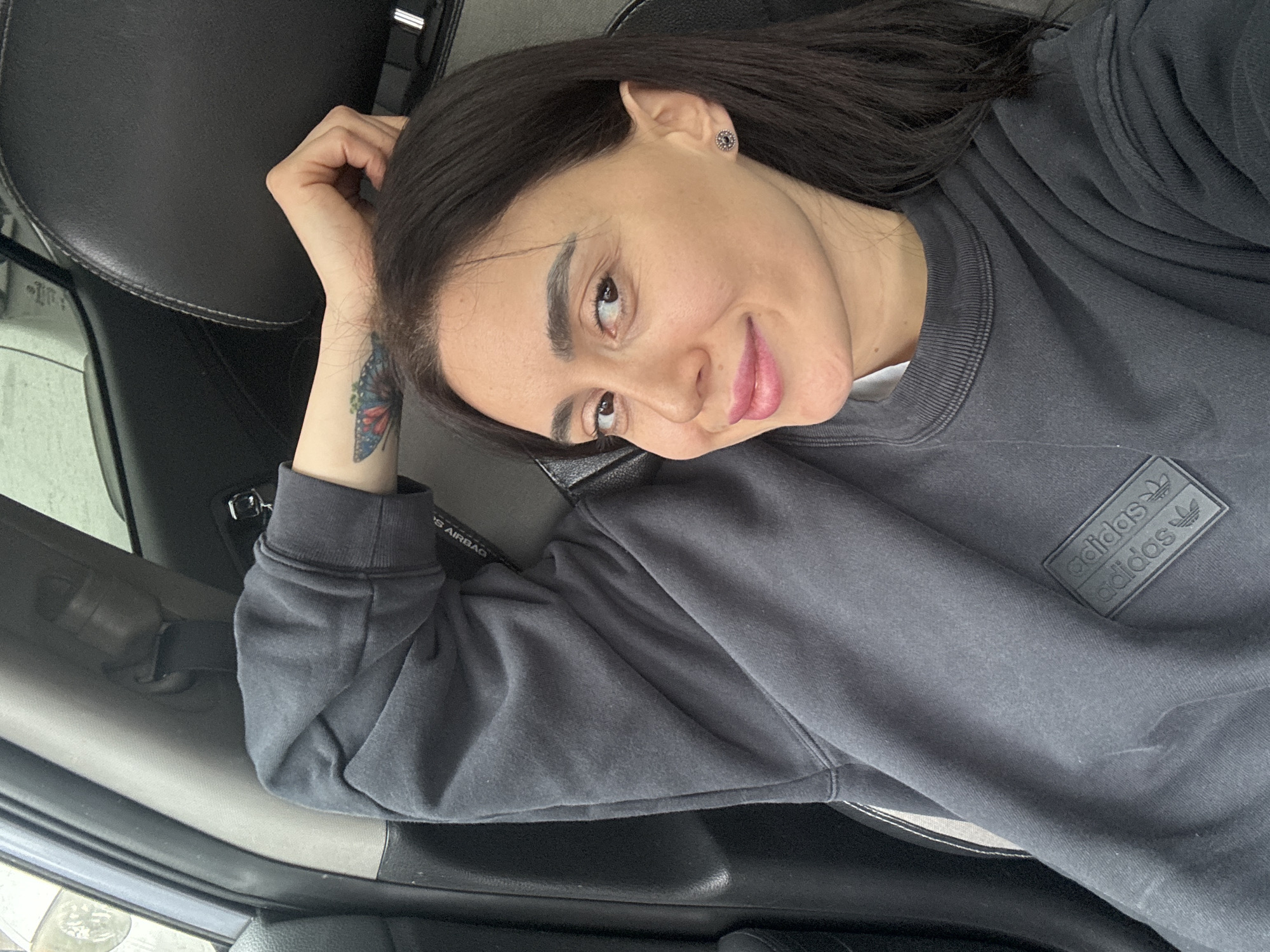 A forensic description of the image reveals a young adult female, appearing to be in her late 20s to early 30s, seated in the passenger seat of a vehicle. Her dark hair is parted on the left side, extending to her shoulders. She has dark eyes, distinct eyebrows, and her lips are tinted with light pink lipstick. A small, circular, dark-colored stud earring is visible in her right ear. She is wearing a dark gray crew-neck sweatshirt featuring a black rectangular patch with two "adidas" logos and trefoil symbols on the left chest/sleeve area. Her left arm is raised, with her hand resting on the back of her head, just above her right ear. A multicolored butterfly tattoo, primarily blue and red with green accents, is visible on her left inner forearm/wrist.
The interior of the vehicle consists of a black leather car seat with a headrest behind her. To her left, a light gray pillar for the seatbelt mechanism is visible, alongside the upper portion of a car door panel. A blurry exterior view, possibly of another vehicle or structure, is visible through the window on the left. On the right side of the frame, near the headrest, a chrome door handle is partially visible. Text indicating "S AIRBAG" is present on the passenger side dashboard or A-pillar. The lighting suggests an overcast or indoor environment. The location of this scene is Tbilisi, Georgia.