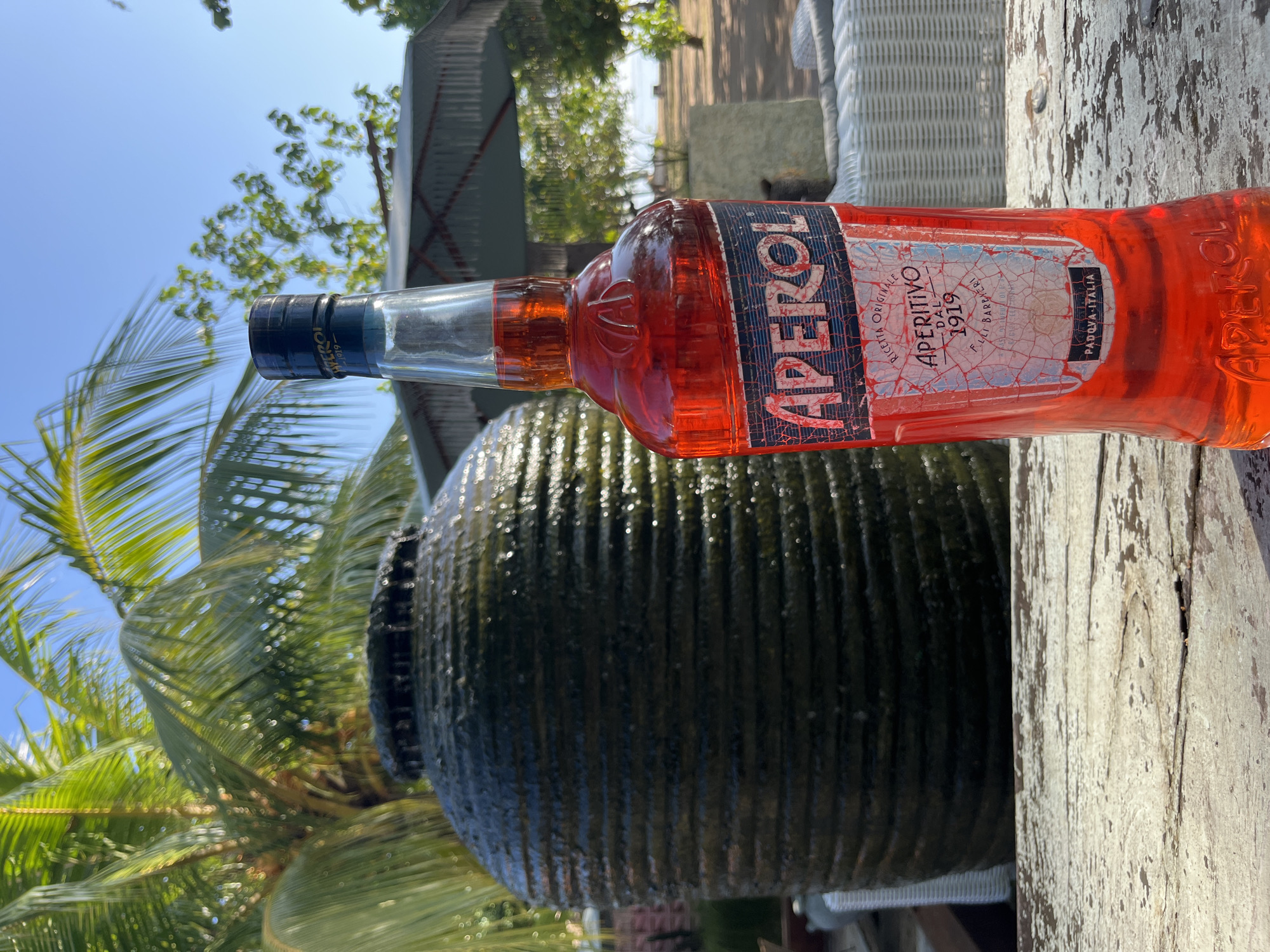 An Aperol bottle stands on a weathered wooden table outdoors, with a blurred tropical background featuring palm trees, a dark, ribbed fountain, and a distant view of the ocean under a clear blue sky. The bottle's label is prominently displayed, showing the brand name "APEROL" in large white letters on a dark blue background, with text indicating "RICETTA ORIGINALE APERITIVO DAL 1919" and "PADOVA - ITALIA" below. The setting appears to be a resort or restaurant area in Kuta Selatan, Indonesia, suggested by the palm trees and beach view. There are no people visible in the image.