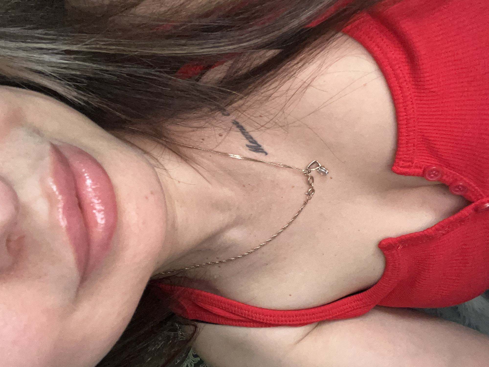 A close-up image captures the upper torso of a person, from the chin to the upper chest. The subject has full, glossy, light pink lips. Their fair to medium complexion skin shows minor texture and freckles/beauty marks, with a slight reddish tone near the philtrum. Dark brown hair with lighter highlights is visible on the right side of the frame. The subject wears a bright red, ribbed top, possibly a tank top or camisole, featuring small red buttons down the front. A delicate gold-colored chain necklace with a small, clear or silver-toned pendant of interlocked shapes rests on the chest. A dark-inked tattoo in cursive script, partially obscured by hair, is visible on the upper right chest/collarbone area. The background is out of focus and indistinct, appearing as a soft, dark surface. The location is indeterminable from the image.