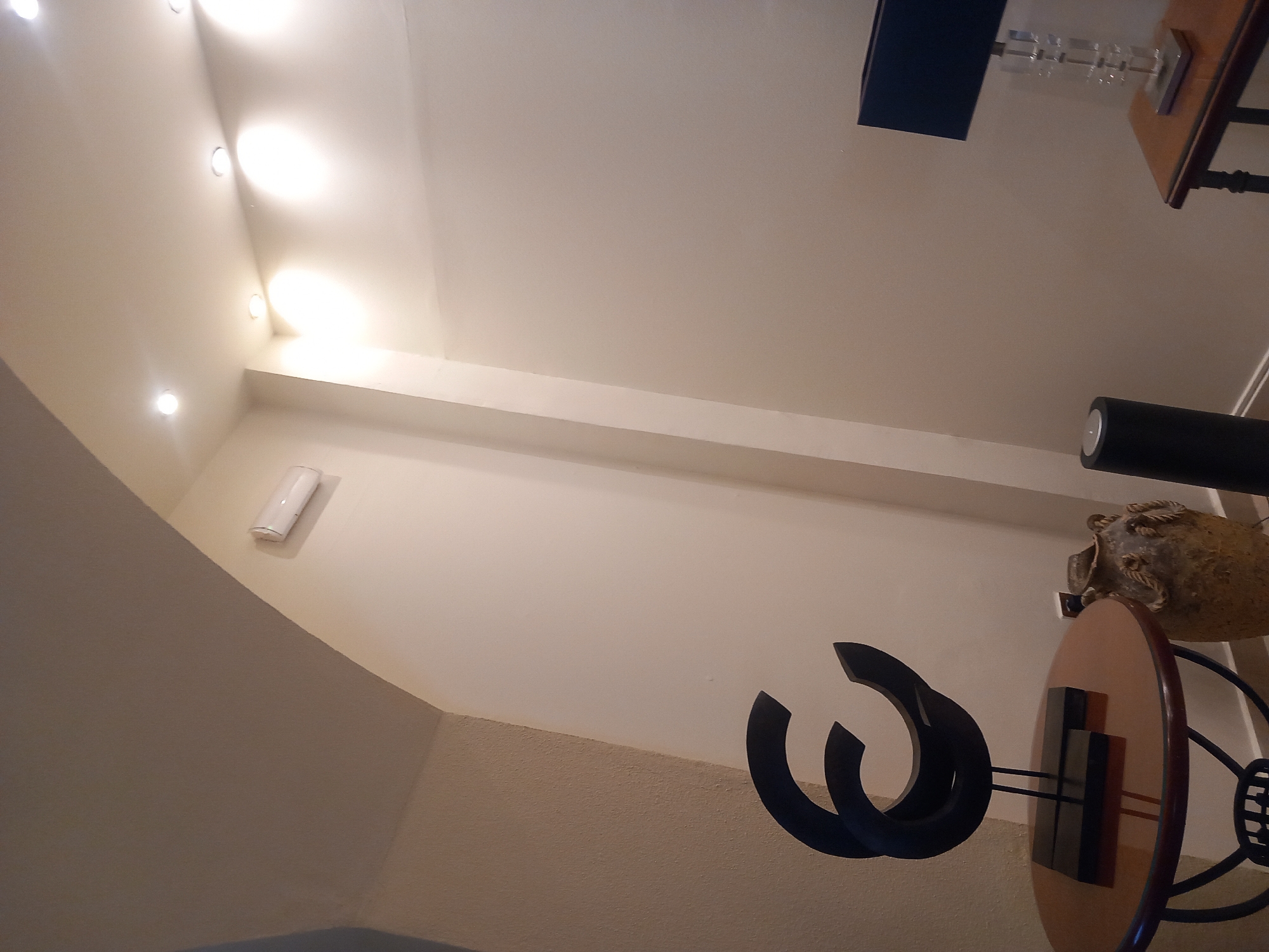 The image depicts an indoor scene within a building, likely a residence or commercial establishment, located in the city of Kos, Greece. The environment appears to be a hallway or an entryway, characterized by curved archways in the ceiling and neutral-toned walls with a textured finish.
Prominently featured is a dark, abstract sculpture with curved elements, positioned on a circular wooden table with a dark rim and a metal base. To the right of the table, a textured, urn-like decorative vase with rope detailing sits on the floor. Further to the right, a cylindrical black object, possibly a speaker or a stand, is visible. In the background, a rectangular table with a wooden top supports a table lamp with a black shade and a clear, faceted base. The lighting in the room is provided by recessed ceiling lights and the table lamp, casting a warm glow on the surfaces. The overall impression is one of modern interior design with a touch of artistic decoration.