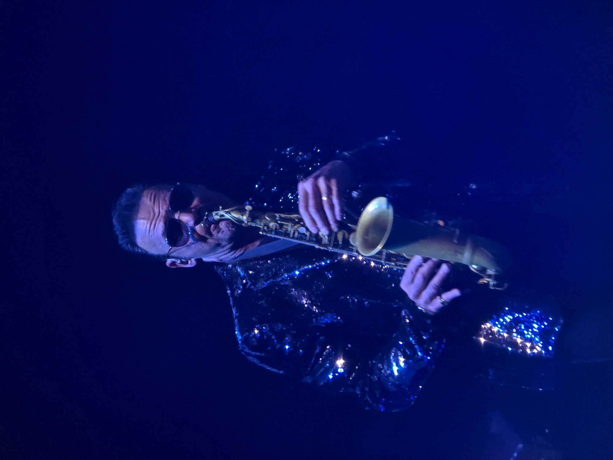 A man in a sequined jacket plays a saxophone on a dark stage. He is wearing sunglasses. The lighting is blue.