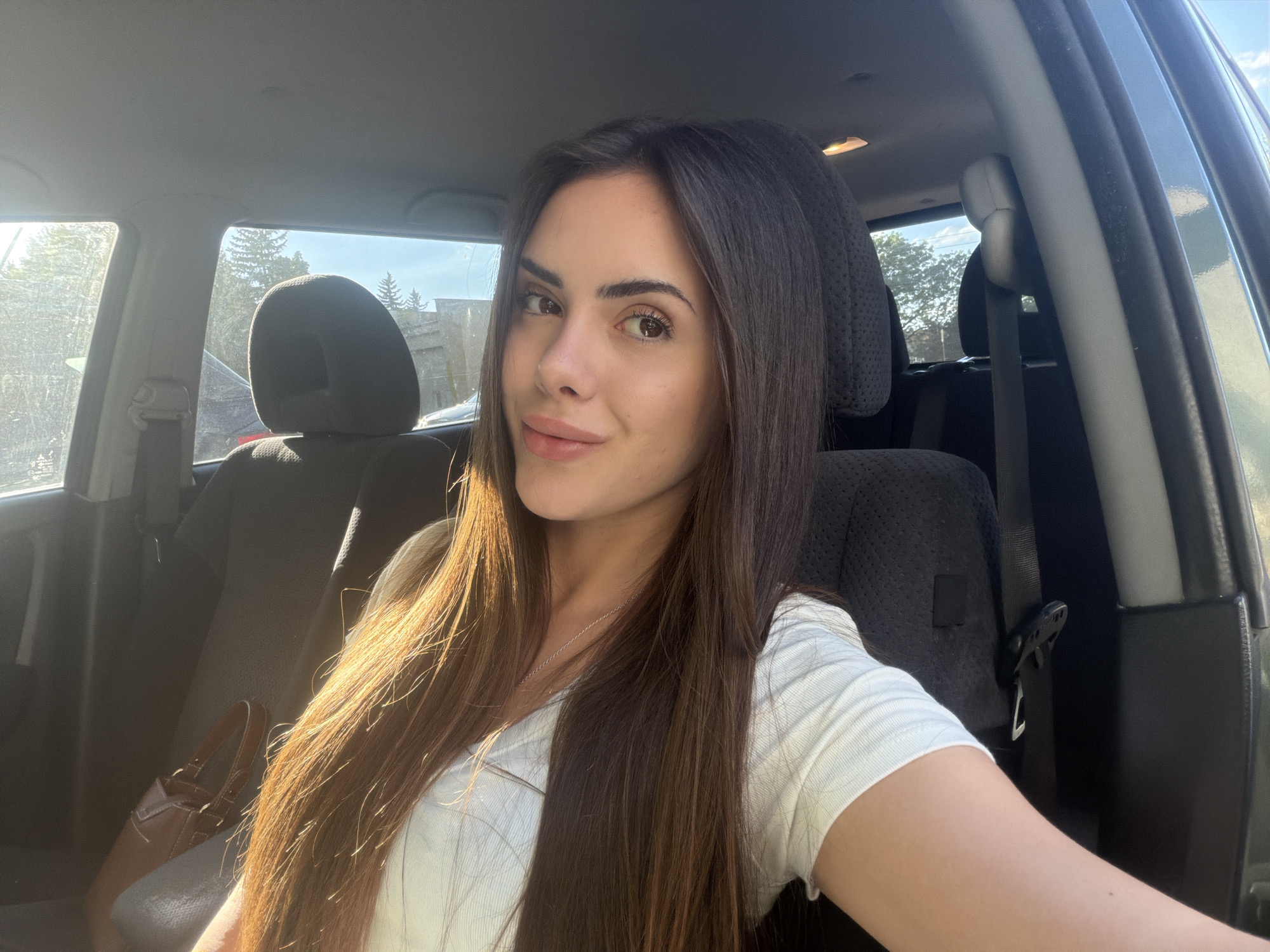 A woman is in the driver's seat of a car. She is wearing a white T-shirt and has long brown hair. The car's interior is visible, including the seats, seatbelts, and dashboard. The scene appears to be outside, with trees and buildings visible through the car windows. The location is not evident from this image.