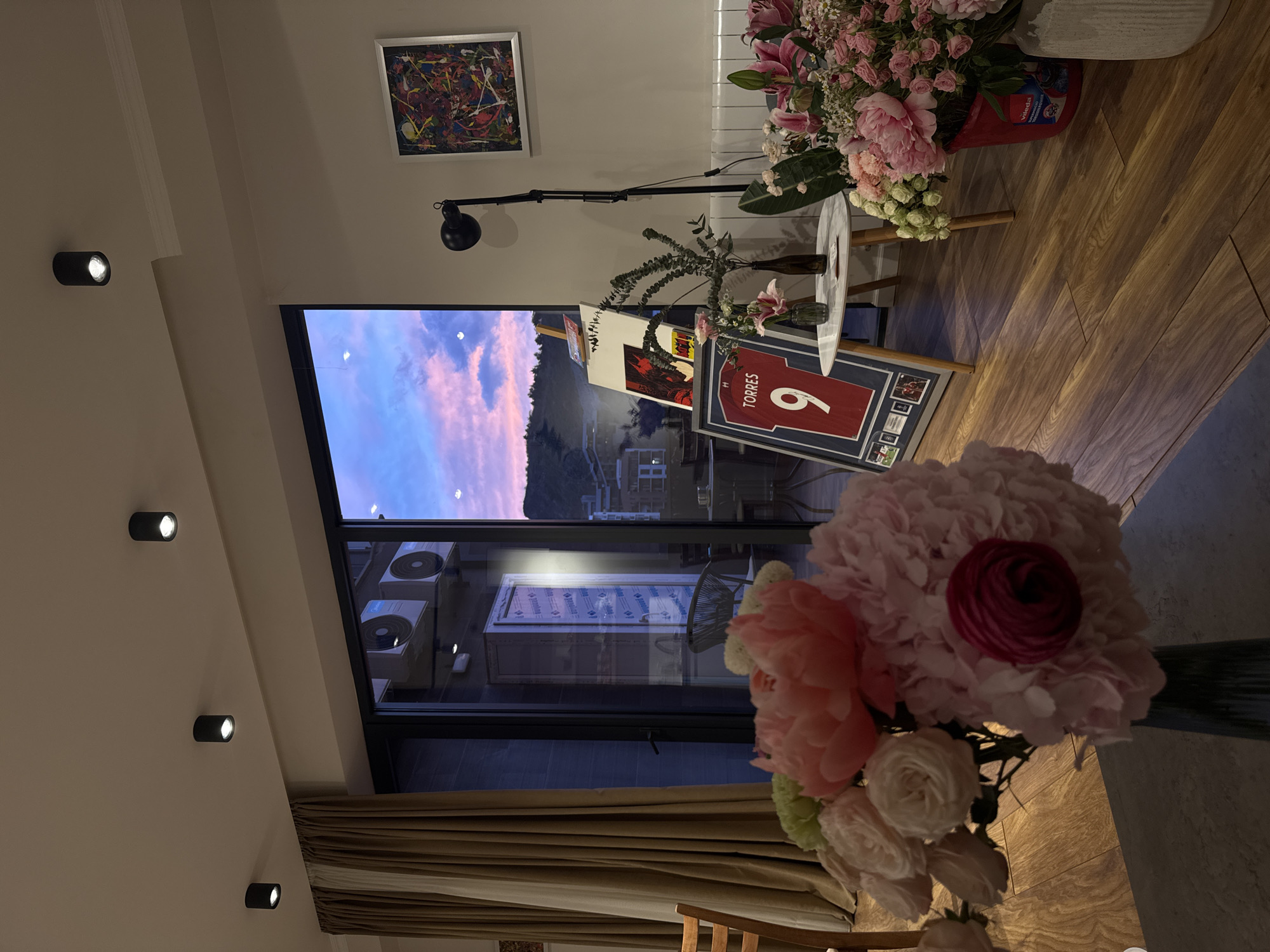 This image captures an indoor living space during dusk or sunset, as indicated by the soft pink and blue hues in the sky visible through a large window. The room features warm wooden flooring and light-colored walls, illuminated by several black cylindrical ceiling spotlights and a floor lamp.
In the foreground, a large bouquet of pink, white, and light green flowers, including prominent peonies and roses, is partially visible. Further into the room, another substantial floral arrangement, featuring pink lilies and other blooms, rests in a red bucket on a small white circular side table. The bucket has "Vileda" text printed on it. On the floor beside this table, a framed red football jersey with the name "TORRES" and the number "9" is displayed. Several other framed items, including what appears to be a red and black print and small photographs, are also visible in the vicinity of the jersey.
On the wall above the floor lamp, a framed abstract painting with splashes of various colors adds a vibrant touch. Through the large window, distant hills or mountains are silhouetted against the colorful sky. Closer to the building, outdoor air conditioning units are reflected in the windowpane, along with text "santina" or "santana" on what appears to be a window frame or reflective surface. A beige curtain is partially drawn at the bottom of the window. The overall scene suggests a cozy and decorated home environment, possibly set for an occasion or simply well-maintained for a relaxing evening. No individuals are present in the image.