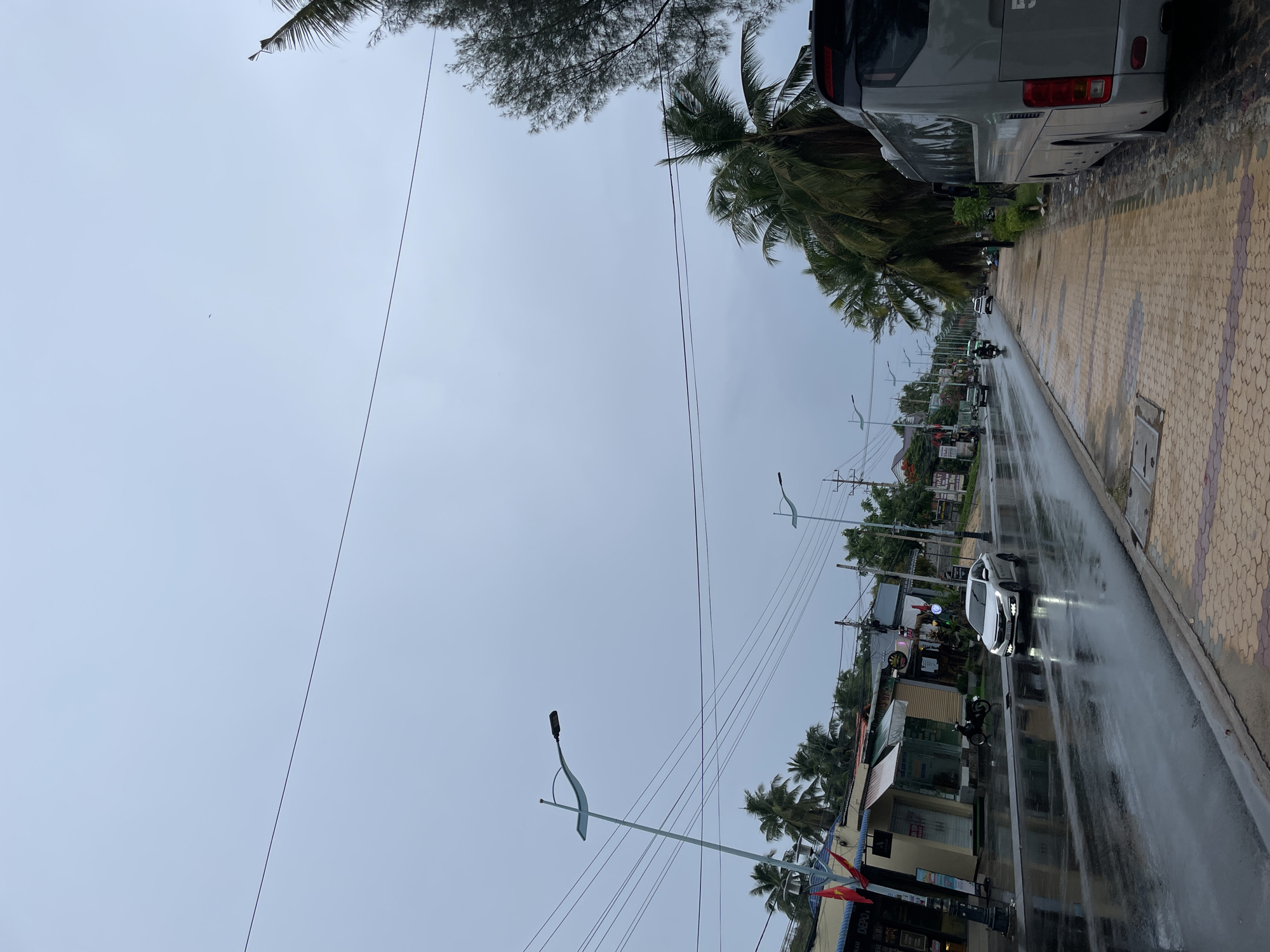 The scene is Phu Thuy Ward, Vietnam, during or shortly after a rainstorm. The pavement and road are wet, reflecting light. A large gray bus is parked along the right side of the road, next to a pedestrian walkway paved in patterned tiles. A white car with headlights on is traveling on the wet road. A scooter is parked on the roadside near a building. Palm trees and other foliage line the street. Power lines are strung above. The sky is overcast. Buildings and storefronts are visible along the roadside. Streetlights are present.