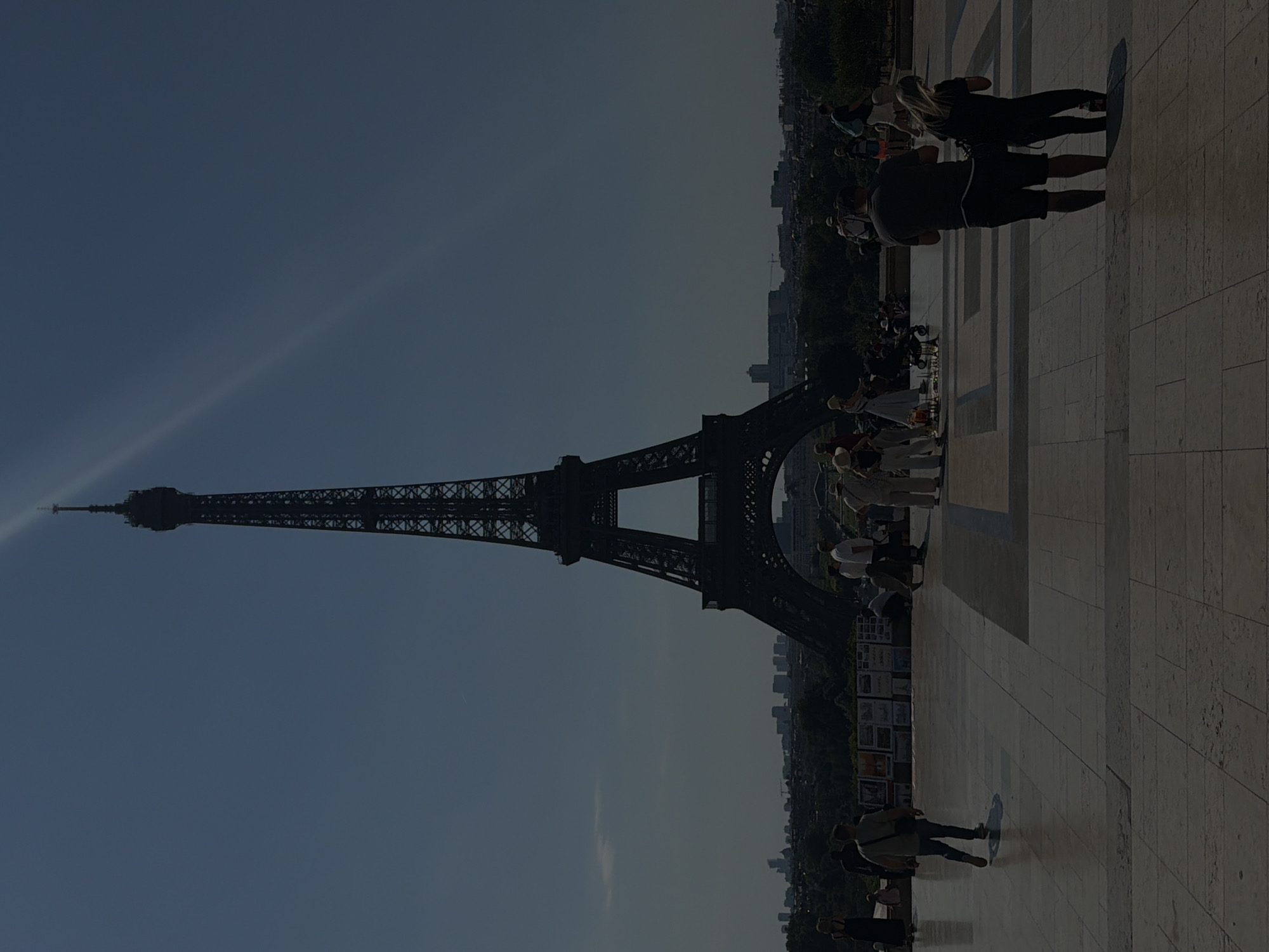 The image shows the Eiffel Tower in Paris, France, on a sunny day. The tower is silhouetted against a clear blue sky, with a few faint clouds visible. In the foreground, a paved plaza is dotted with people walking and taking pictures. Some people are gathered around a vendor selling miniature Eiffel Tower replicas. A man and a woman in the foreground are looking at the Eiffel Tower. The atmosphere appears relaxed and tourist-oriented.