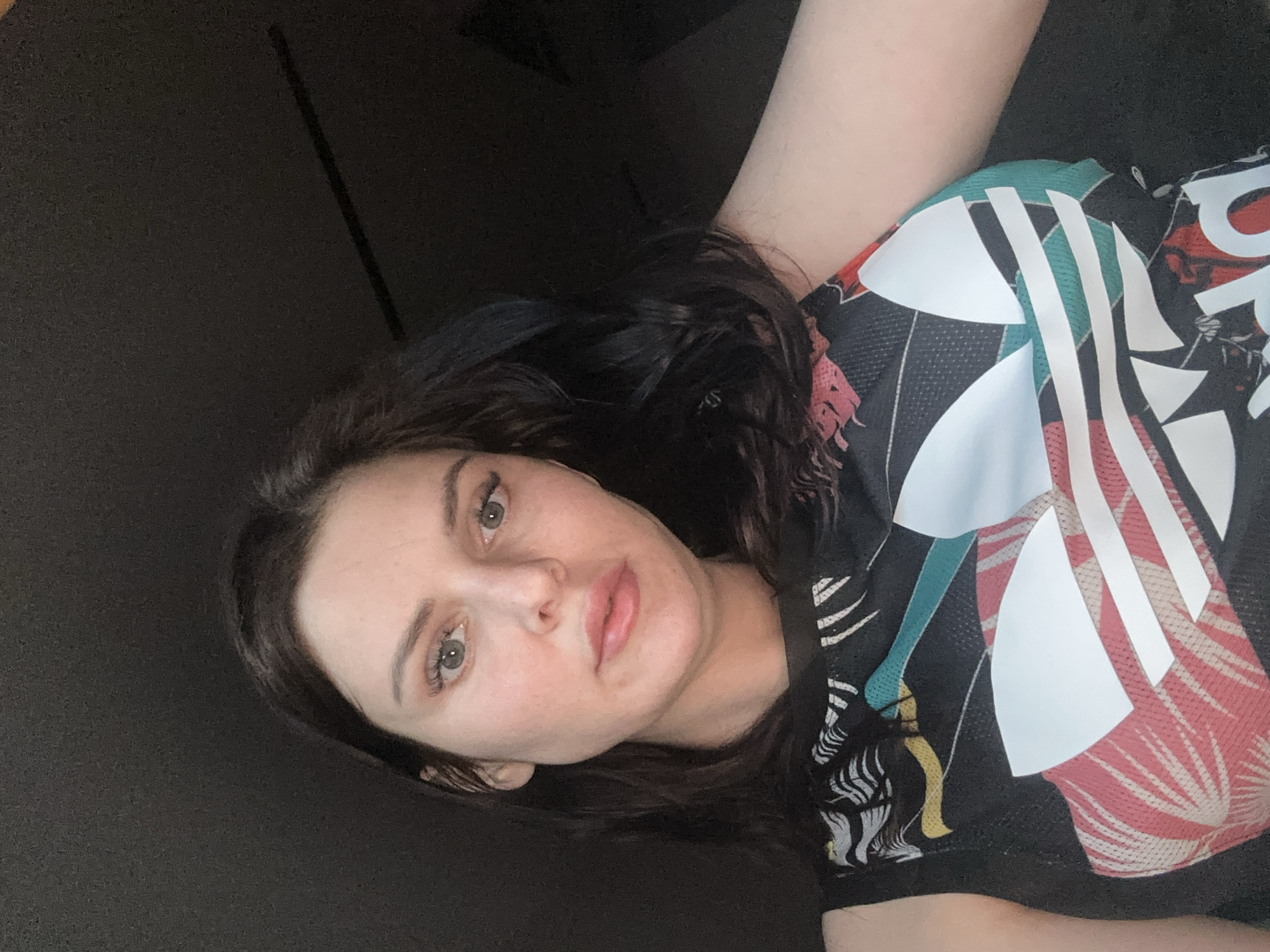 A woman with dark hair and light-colored eyes is shown in a close-up selfie. She is wearing a black sleeveless top with a colorful, abstract design featuring white Adidas stripes. Her expression is neutral, and she is looking directly at the camera. The background is dark and indistinct, suggesting an indoor setting with low lighting.