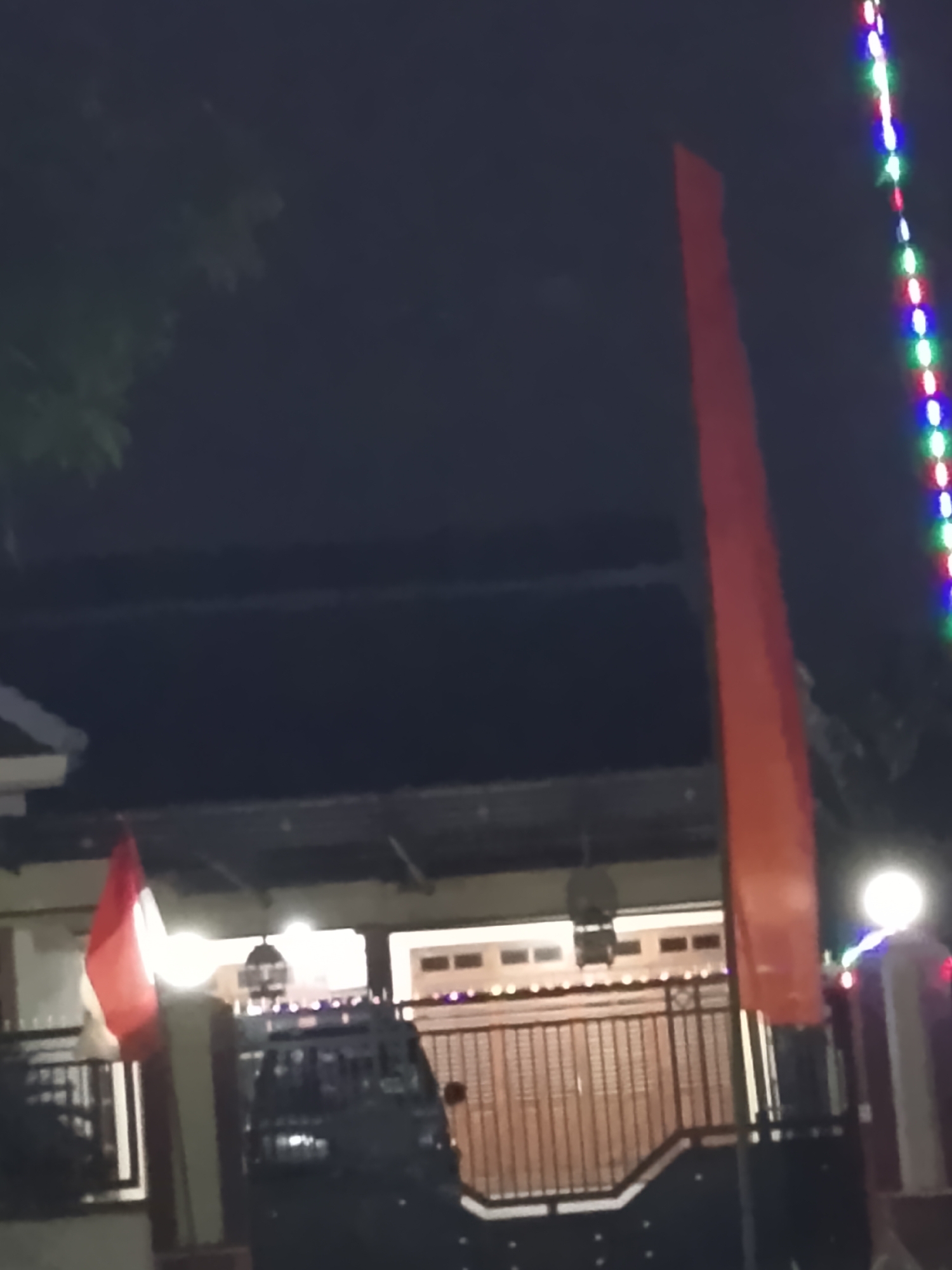 A dimly lit, exterior shot captures a residential scene at night. A red and white flag, possibly the Indonesian flag, is prominently displayed on the left side of the frame, attached to a pole. To the right, a tall, thin, red flag, likely a banner or decorative item, stands unfurled. A string of colorful LED lights, alternating between white, green, blue, and red, hangs vertically on the far right. In the background, a dark-colored vehicle, possibly a van or truck, is parked behind a black metal fence. The house behind the fence has a dark roof and illuminated windows. The overall impression is of a festive or celebratory nighttime setting in a residential area of Kelurahan, Indonesia, given the presence of flags and decorative lights.
