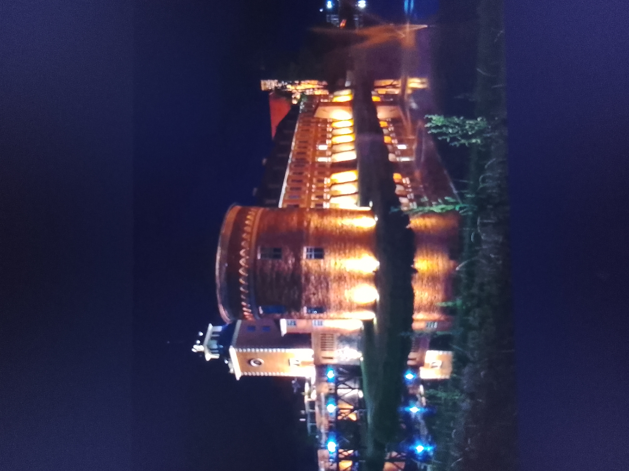 The image depicts a night scene of a brick building, possibly a historical structure or a themed attraction, illuminated by warm yellow and blue lights. The building has a cylindrical tower and appears to be multi-story with arched windows. Some greenery, likely trees or bushes, is visible to the right of the structure. The overall impression is of a well-lit, possibly festive, nighttime environment. The image is oriented sideways, requiring rotation for proper viewing. The location specified is the city of Prisel'e, Russia.