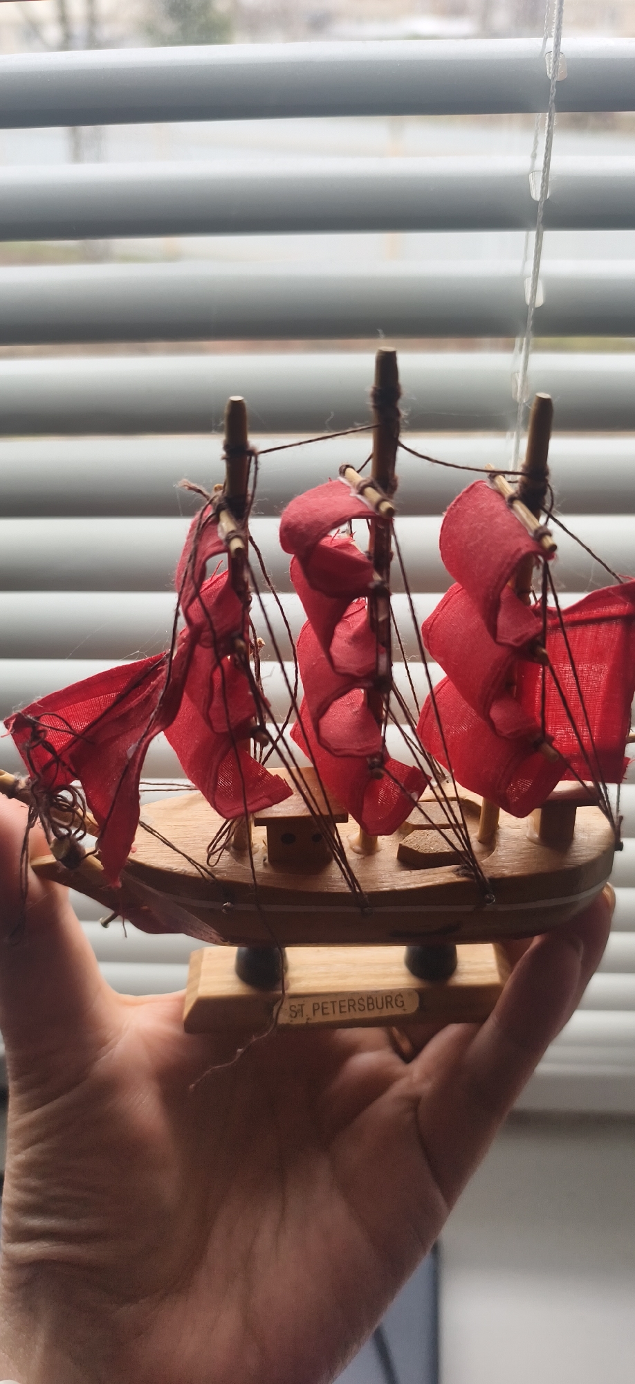 A person is holding a small wooden model of a sailboat with red sails. The model has "ST. PETERSBURG" printed on its base. The background shows white blinds. The location is not Фоки, Russia, but potentially St. Petersburg based on the text on the base of the model.