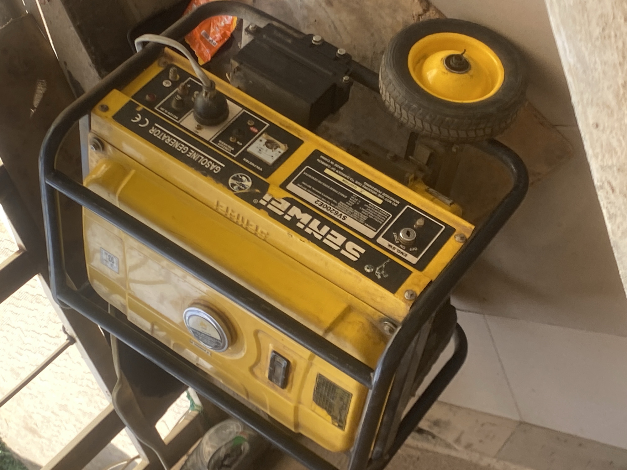 The image shows a yellow SENWER SV6200E2 gasoline generator within a black metal frame. The generator's control panel is visible with labels like "GASOLINE GENERATOR CE," "ENG.SW," "OFF ON START," "VOLUMETER," and "CAUTION." A black component is mounted on top of the generator, and a yellow-rimmed wheel is situated to the right. The generator is in an indoor setting, possibly a storage area or garage. The location is not possible to determine based on the image.