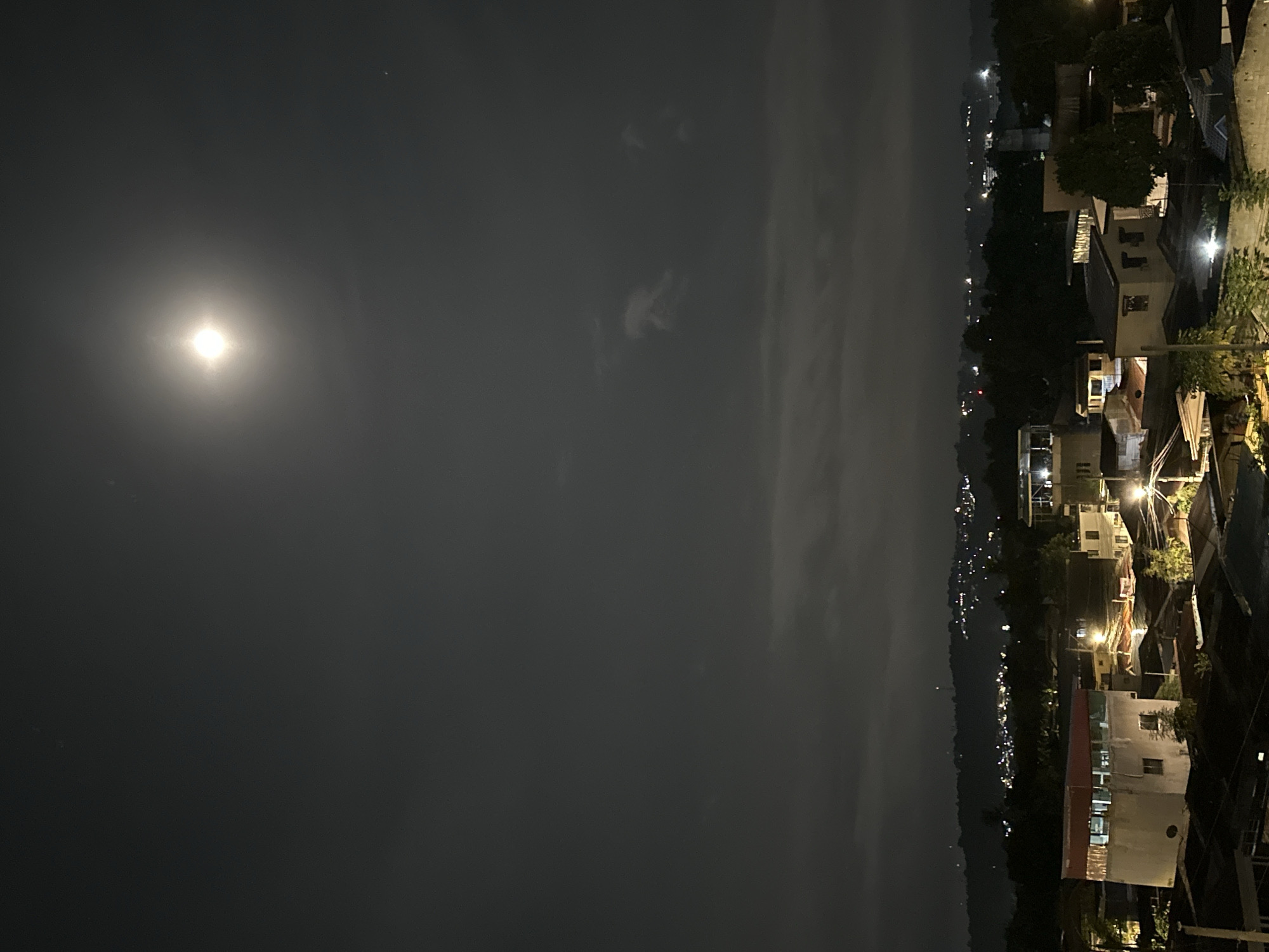 A night scene in Davao City, Philippines, features a view of buildings and city lights against a dark sky with a bright moon. The buildings are situated on elevated terrain, with lights scattered throughout the area. The foreground is obscured by darkness, allowing the city lights and the moon to stand out. The sky is overcast, though the moon is still visible.