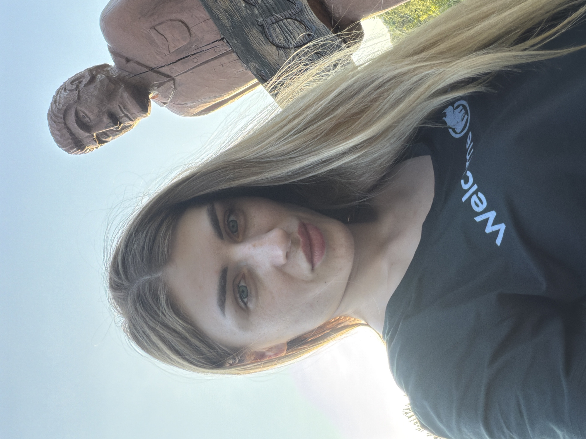 A woman with blonde hair and green eyes takes a selfie outdoors. She is wearing a dark green t-shirt with white text and a logo. Behind her, in the background, is a wooden statue of a man. The sky is clear and blue. The lighting suggests it is late afternoon or early evening.