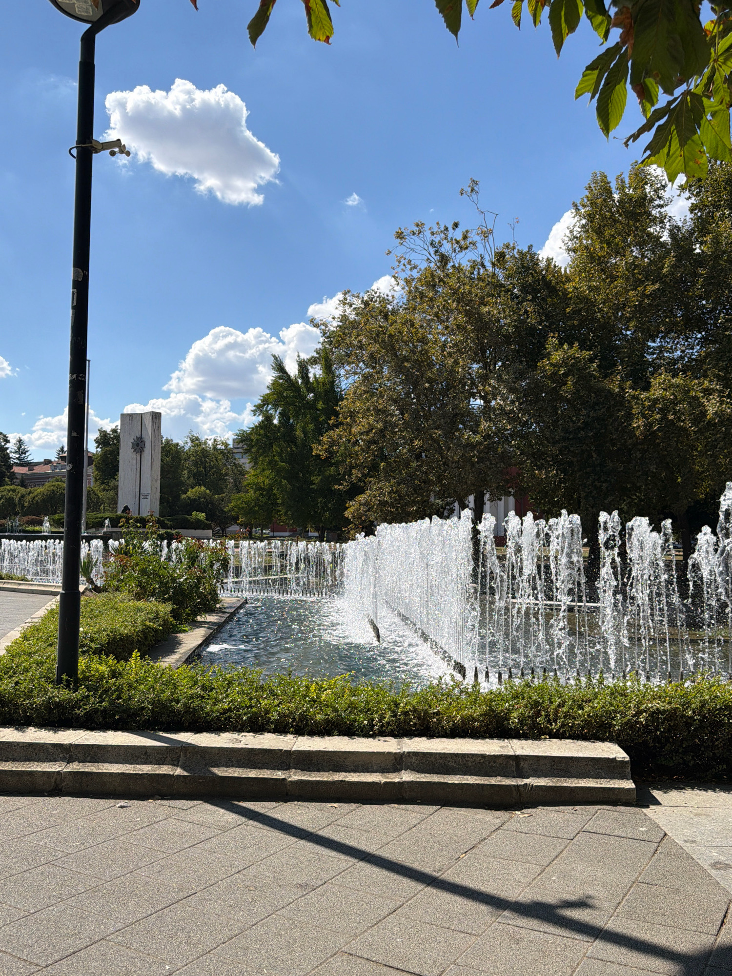 The image depicts a public park with a prominent fountain in the foreground, featuring multiple jets of water spraying upwards. The fountain is bordered by manicured green bushes. In the background, a tall, rectangular monument stands against a backdrop of lush trees and a blue sky with scattered white clouds. A lamppost is visible to the left of the fountain, with what appears to be a weather sensor attached. The ground in front of the fountain is paved with grey stone tiles, and there are stone steps leading up to the fountain area. The scene suggests a sunny day in a well-maintained urban park. This is the general area of the Monument of Bulgarian-Soviet Friendship in Pleven, Bulgaria. There are no people or animals visible in the image.