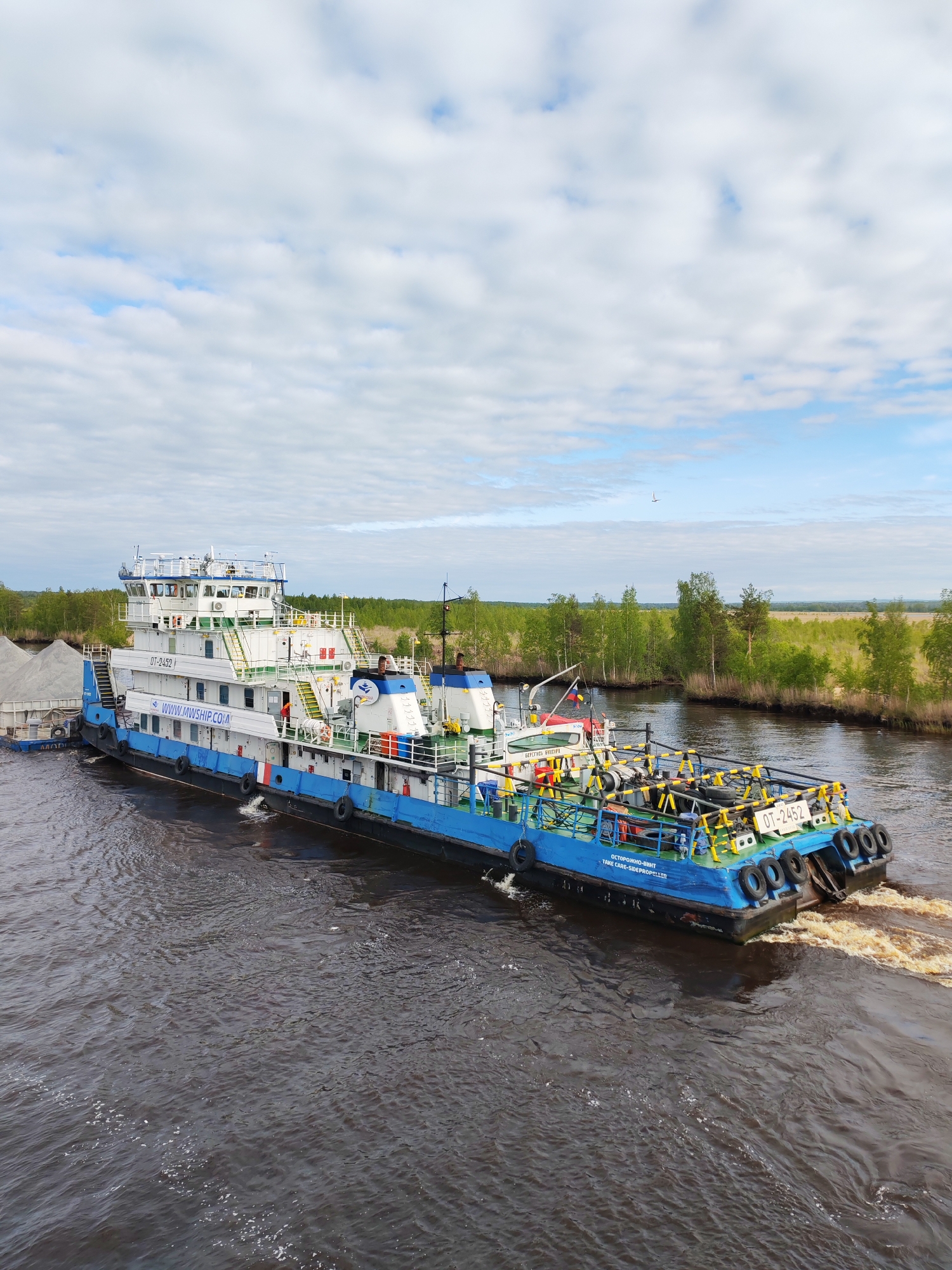 The image shows a blue and white river tugboat, identified as "OT-2452," with a barge attached to its side. The vessel is moving through a dark, possibly muddy river in Ozernoe Ust'e, Russia, as evidenced by the vessel's official website. The barge is loaded with gray material, possibly gravel or sand. The tugboat has multiple decks and a control tower. The stern of the tugboat shows a warning message, "ОСТОРОЖНО-ВИНТ TAKE CARE-SIDEPROPELLER." The river is bordered by green trees and vegetation. The sky above is partly cloudy with patches of blue.