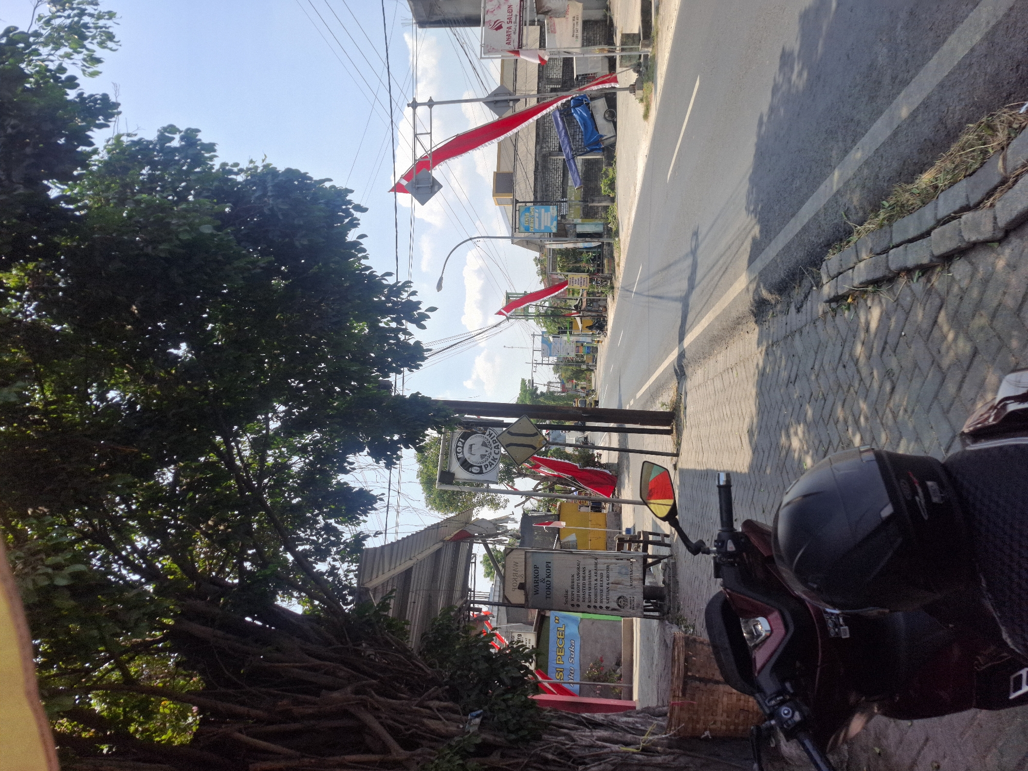 This is a street-level, eye-level, eye-level photograph taken in Karanggeneng, Indonesia, on a bright, sunny day. The scene is dominated by a paved road with a white lane marker, bordered by a sidewalk constructed of brick pavers. A large tree with thick, gnarled roots shades a portion of the left side of the image, its branches extending over the sidewalk and a small awning.

Along the sidewalk, a motorcycle is parked, facing away from the camera, with a black helmet resting on its seat. A woven basket is also visible near the tree. Further down the sidewalk, a warung (small shop) with a metal awning is set up, featuring a sign for "WARKOP & TOKO KOPI" which lists various coffee and snack options. Several red and white Indonesian flags are displayed, some hanging from poles and others attached to a street light.

Across the road, various buildings are visible, including a structure with a brick facade and a blue tarp. A sign for "ANATA SALON" is prominent on the right side of the street. Power lines crisscross the sky. The road itself appears to be a thoroughfare, with evidence of traffic including shadows cast by passing vehicles. The overall atmosphere suggests a typical street scene in a Indonesian town on a clear day.