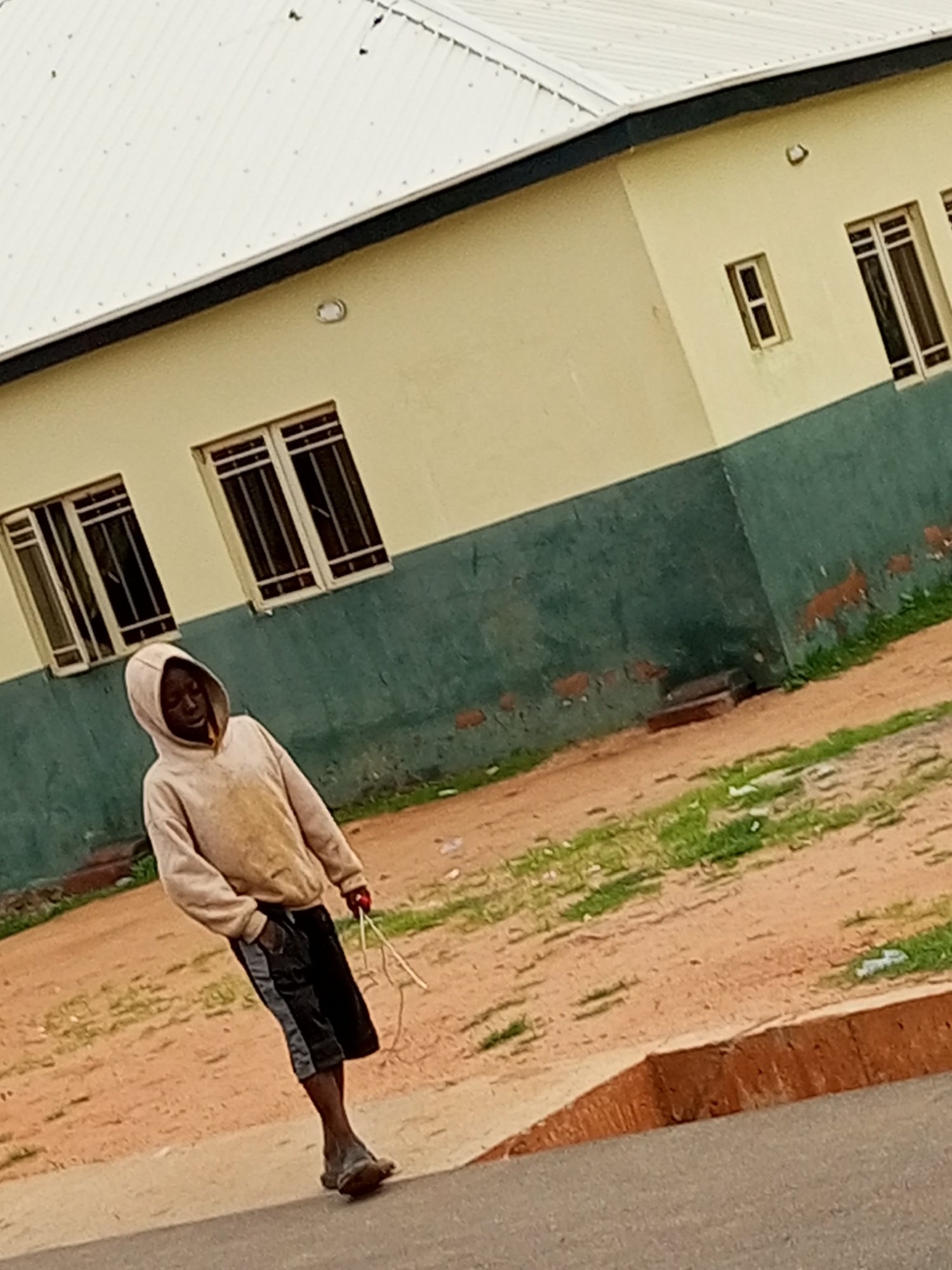 A young boy wearing a light-colored hoodie and black shorts stands on a dirt path beside a building in Tempure, Nigeria. The boy is looking down, and his hands are by his side. He is holding some white string in his right hand. The building has a light yellow facade on its upper portion and a dark green facade on its lower portion. There are several windows on the building, some with bars. The ground is covered in dirt and patches of grass. The scene appears to be an outdoor environment.